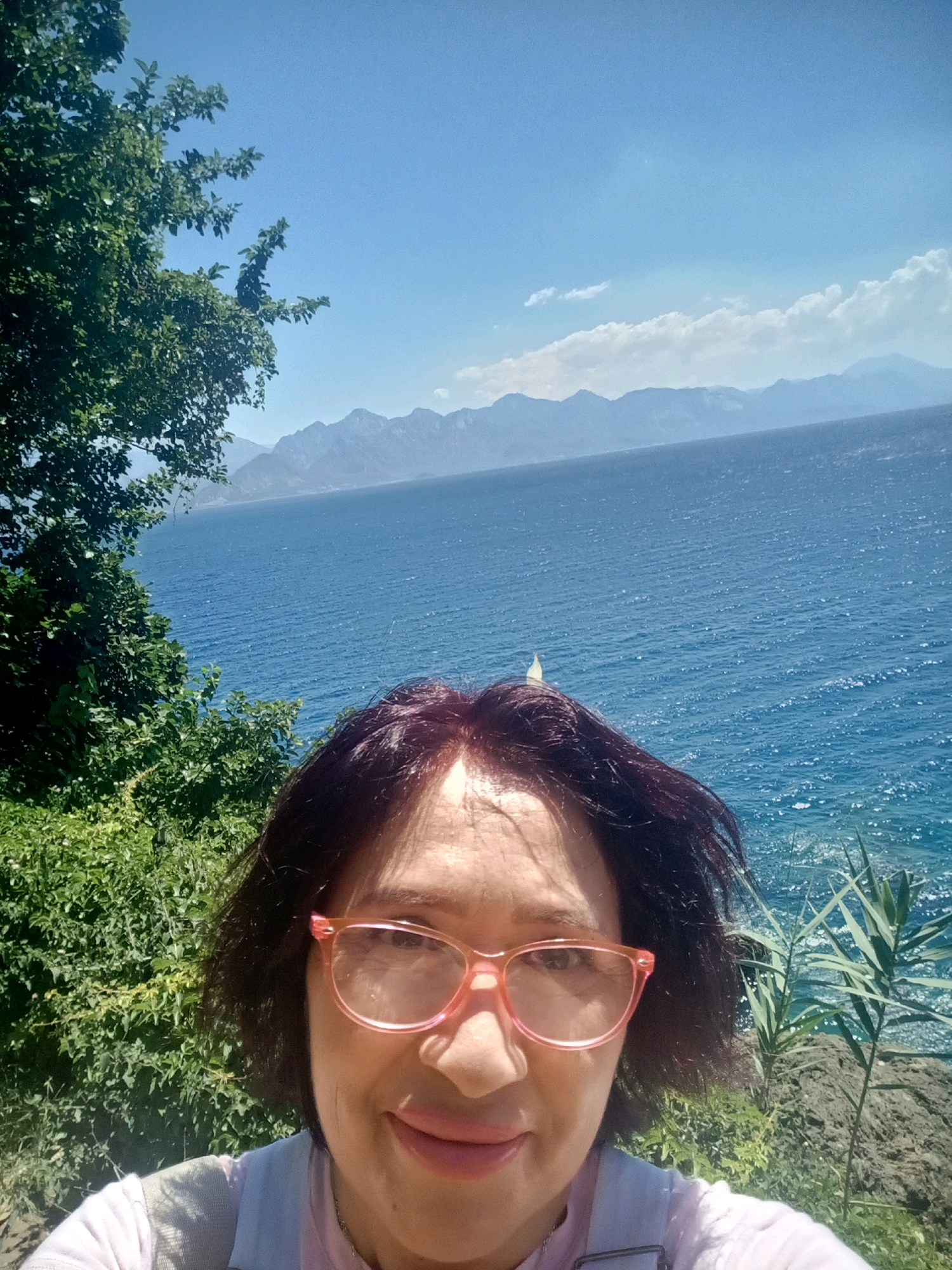 A selfie is taken outdoors with a woman in the foreground. She has short, dark hair and is wearing pink-rimmed glasses and a light pink shirt. She is smiling, and her face is lit by sunlight, creating highlights on her forehead.
The background of the image features a bright blue sea under a clear sky. In the distance, a range of mountains can be seen, with some clouds near the horizon. Along the coastline, there is lush green foliage and some rocky terrain. A sailboat is visible on the water. The scene suggests a location on the coast of Antalya, Turkey.