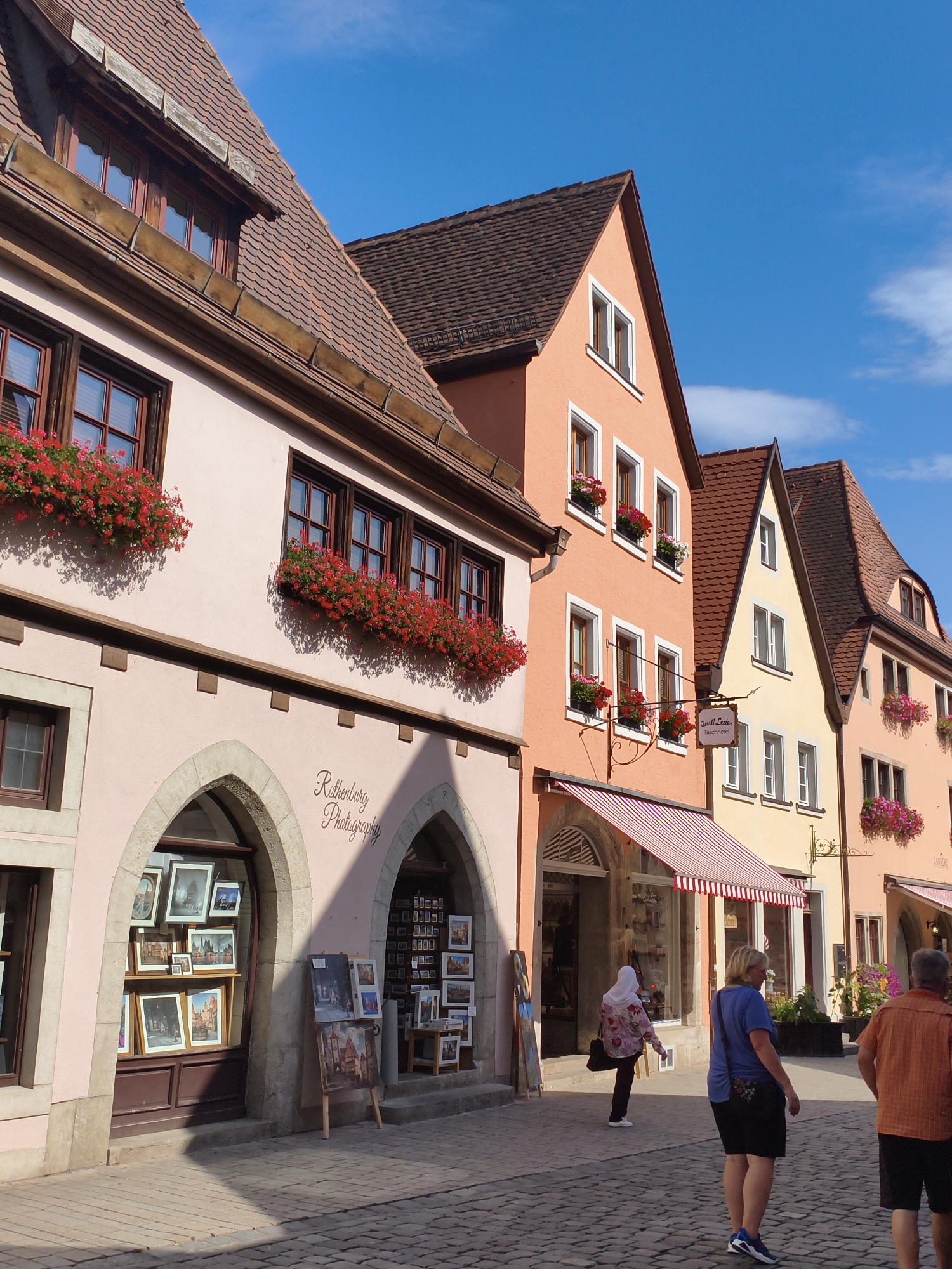 This is a vertical, medium shot photograph of a charming street scene in Rothenburg ob der Tauber, Germany, captured on a bright, sunny day with a clear blue sky. The image is dominated by the facades of historic, colorful buildings lining a cobblestone street.
In the foreground, the cobblestone street is visible, with distinct shadows cast by the buildings, indicating the sun is high in the sky. To the left, a pastel pink building with a terra cotta tiled roof features several windows adorned with vibrant red flowers in planters. The lower level of this building has an arched entryway with stone detailing, displaying framed pictures and artwork within. A sign above this entryway reads "Rothenburg Photography" in a cursive font. Another similar arched entrance is visible further down this building, also displaying artwork.
Further to the right, a row of buildings with peach, yellow, and other pastel-colored facades are lined up. These buildings also have terra cotta tiled roofs and windows decorated with flowers. Striped awnings extend over some of the shop entrances.
Several people are present in the scene. On the right side of the street, a man wearing an orange shirt and black shorts walks away from the camera. In front of him, a woman wearing a blue shirt and black shorts walks towards the camera. Further back, a person wearing a light-colored head covering and a patterned long-sleeved shirt walks away from the camera. Several other figures are visible in the shop windows and further down the street.
The overall impression is one of a picturesque, well-preserved medieval town, bustling with activity and bathed in warm sunlight.