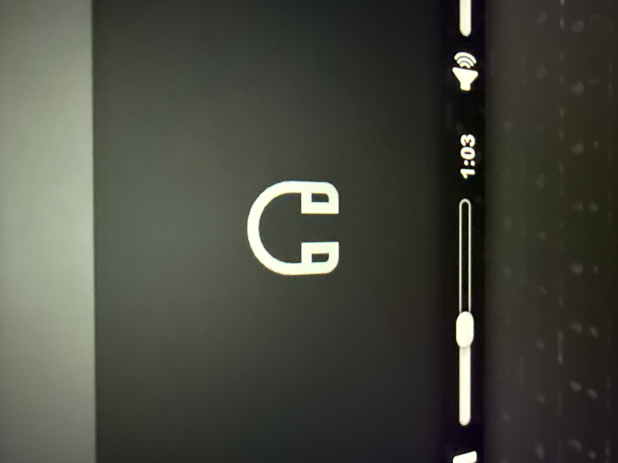 The image displays a close-up, slightly angled view of a digital screen, likely a laptop, showing a media player interface. The scene does not depict or provide any visual information related to the city of Kardzhali, Bulgaria.
Centrally positioned on a uniform dark grey background is a prominent white outline icon of over-ear headphones. Below this icon, a white horizontal progress bar is visible, with a white, oval-shaped scrubber positioned towards the left, indicating media playback progress. To the right of the progress bar, the timestamp "1:03" is displayed in white digits. Further right, a white speaker icon with sound waves emanating is present, signifying active audio. The very bottom of the image shows the blurred, out-of-focus texture of a keyboard. The screen itself exhibits visible pixelation due to the close-up photography. The upper section of the image features a subtle horizontal line, which appears to be the physical top edge of the screen or its bezel, separating the dark display area from a lighter grey surface above it, consistent with a background wall or surface behind the device.