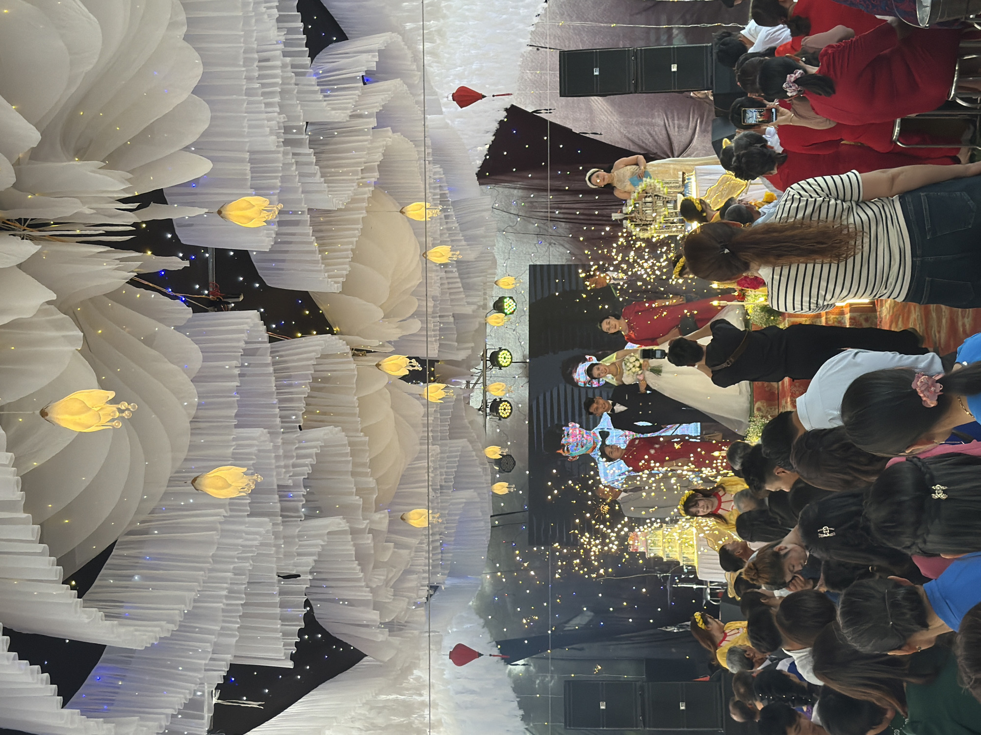The image depicts an indoor wedding event, likely in Bình An, Vietnam. The venue is decorated with large white floral arrangements and strings of lights. A stage is visible with several individuals, possibly the wedding party, dressed in formal attire. Attendees are observing the event; some are taking pictures. Sound equipment is present, indicating audio amplification. The environment suggests a festive and celebratory atmosphere.