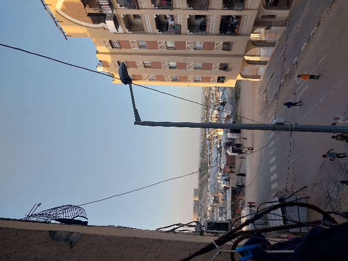 The image captures an elevated view of a street scene in Khan Yunis, Palestinian Territory, under a clear blue sky. On the left, a damaged beige building facade with a broken black mesh is visible, along with a cluster of tangled wires and a blue clothespin. A prominent grey street light pole stands in the center foreground, extending its arm over the road, with multiple wires attached.
The street below is paved with white crosswalk markings. Children are active on the street: three are running towards the bottom left, and two individuals, one in a blue hoodie and one in a yellow hoodie, stand further down the road. Several other people, including adults, are visible walking on the street and congregating near small structures in the mid-distance.
To the right, a multi-story beige building features arched architectural elements, balconies with laundry hanging, and windows with decorative grates. The ground floor has a series of arches, beneath which a red car is partially visible.
In the background, a vast, arid landscape extends, densely populated with numerous white and grey tents and makeshift shelters, indicating a large encampment. Hills or elevated ground are visible on the horizon, topped with structures. The overall environment appears dry and dusty, with a mix of established urban structures and temporary dwellings.
