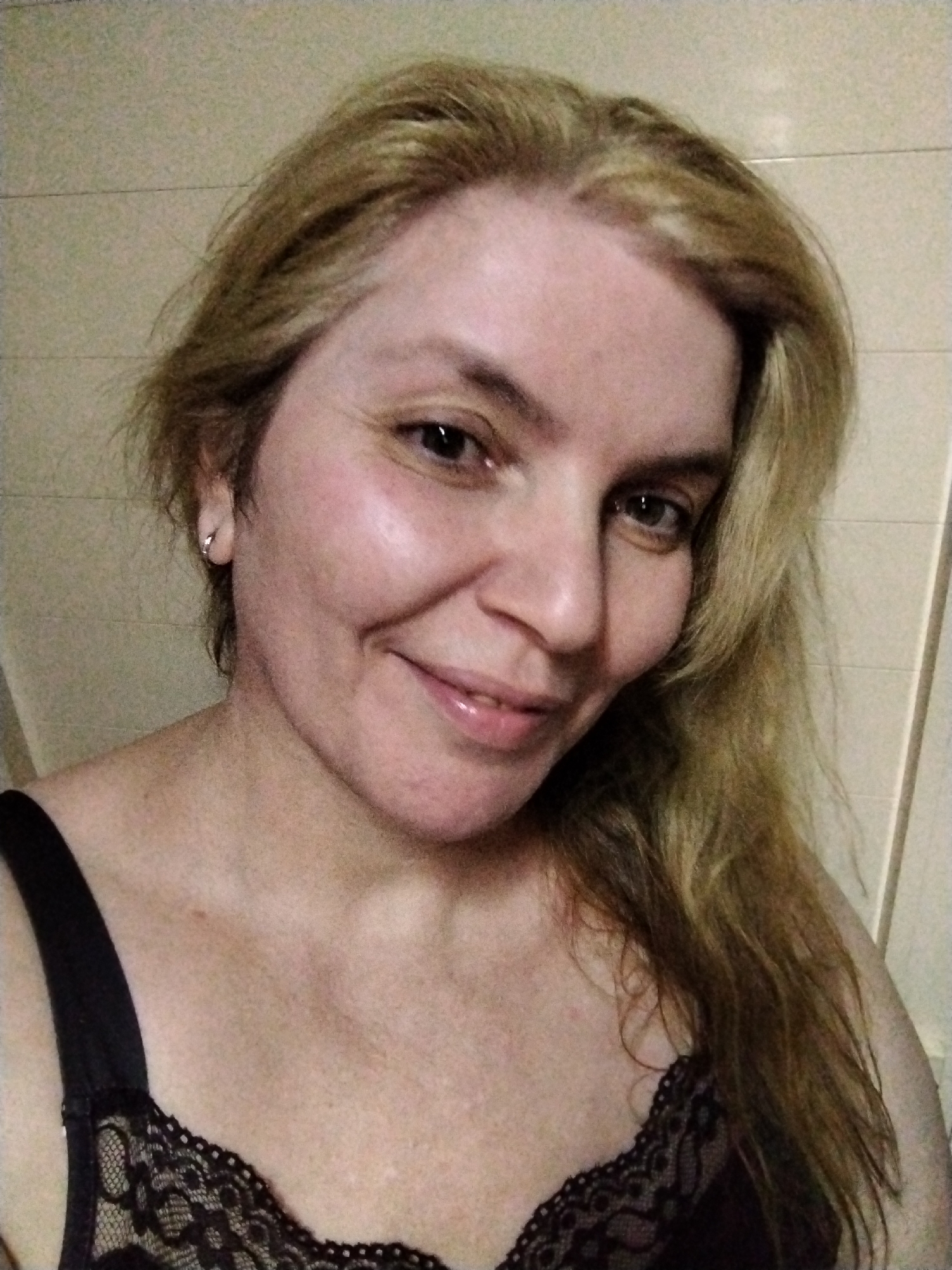 A medium-shot, head-and-shoulders selfie photograph shows a middle-aged white female with fair skin, hazel eyes, and blond, shoulder-length hair. She is wearing a black lace-trimmed camisole. She is smiling slightly. The background consists of a light-colored, horizontally-paneled wall. There is no indication of a specific location within Al Mas'oudi, United Arab Emirates.