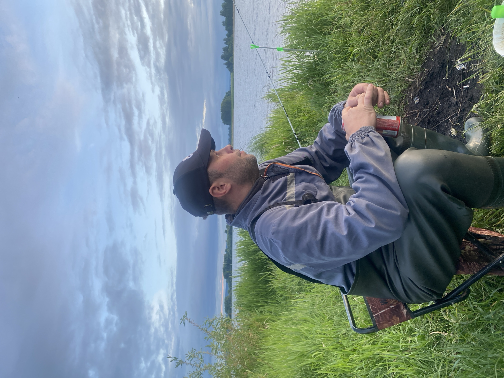 The scene depicts an outdoor setting in Elovo, Russia, on the bank of a body of water, likely a river or large lake, during what appears to be late afternoon or early evening. A Caucasian male, approximately 30-40 years old, is seated on a small, dark-framed folding stool with a camouflage pattern fabric seat. He wears a dark baseball cap, a grey long-sleeved work jacket or shirt, and dark green chest waders with integrated boots. His head is turned to the right, and he gazes attentively towards the water. His hands are clasped together, holding a red aluminum beverage can.
To the man's right, a white fishing rod is positioned, supported by a neon green rod holder planted in the bank. Another similar green rod holder is visible further out in the water, suggesting a fishing setup. The immediate foreground consists of dense, green grass. To the man's right, near his feet, there is a small patch of dark, disturbed earth containing minor debris, including what appears to be several small white fragments, possibly refuse. A small white flower grows near the disturbed earth.
The background features a wide expanse of calm water under a partly cloudy sky, which displays varying shades of light blue and grey. Across the water, a distant shoreline is visible, densely covered with green trees and vegetation, extending to a horizon where faint warm hues are present, characteristic of a setting sun. To the left of the frame, some leafy green branches of a shrub or tree are visible.