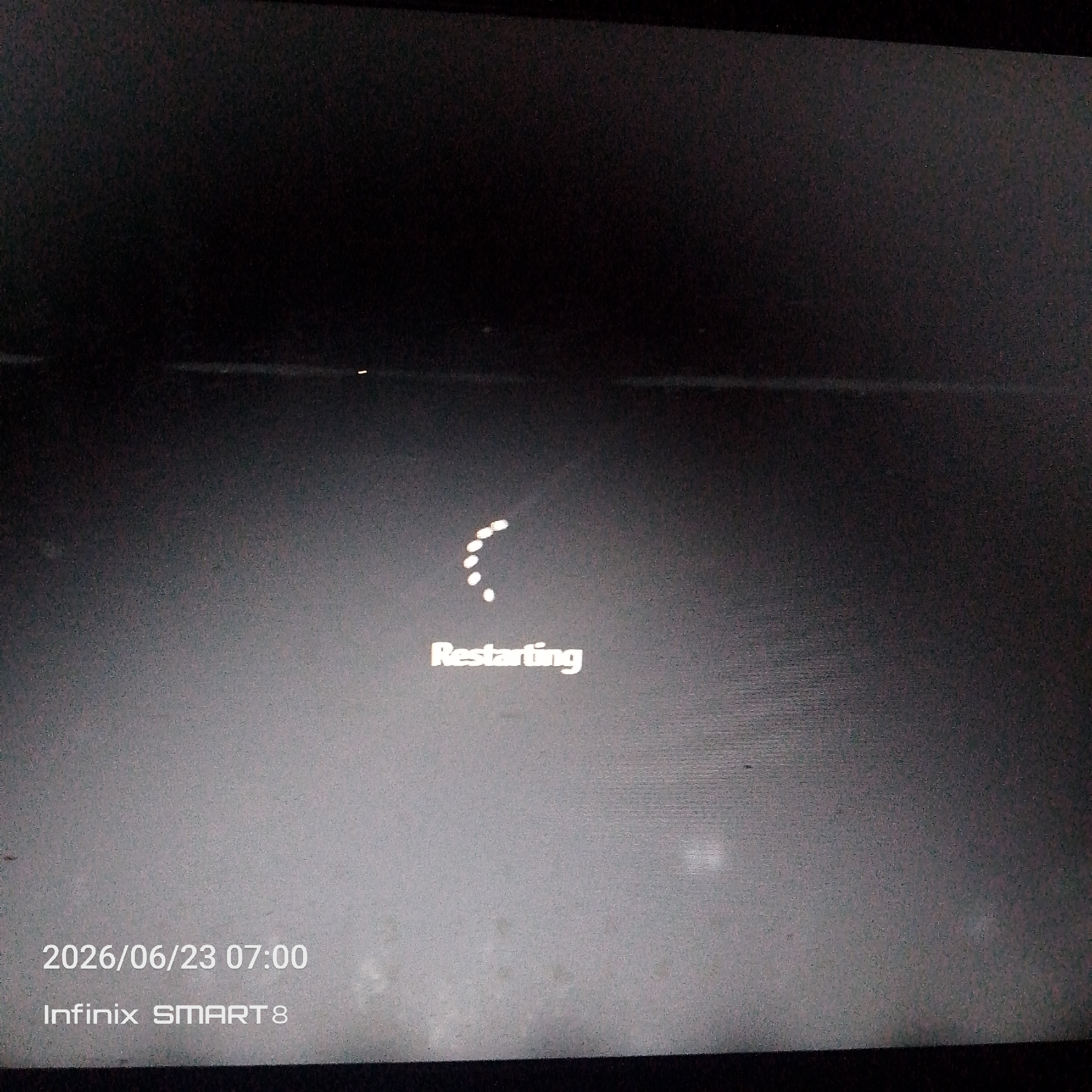 The image is a close-up, slightly blurry photograph of a dark screen displaying a "Restarting" message. A circular animation of dots, indicating the process, is visible above the text. The text "Restarting" is in a white, sans-serif font. Below this, the date "2026/06/23 07:00" and the device name "Infinix SMART 8" are displayed in the same font. The overall lighting is dim, with some faint reflections on the screen. The background is dark and featureless. The image appears to be taken of a device screen, likely a smartphone or tablet, during a restart sequence. There are no people, city elements, or any environmental context visible.