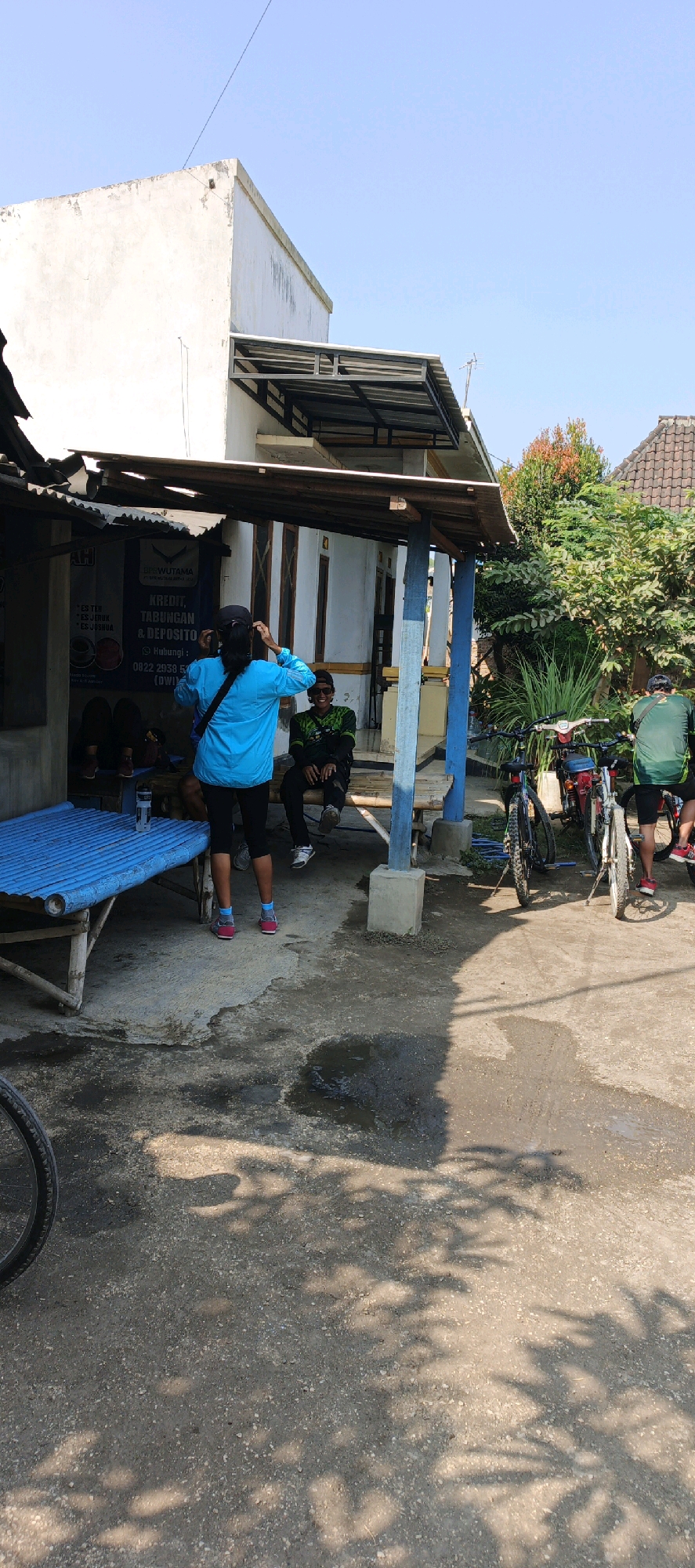 The image depicts a daytime scene outdoors, likely in a rural or semi-urban setting in Mojomulyo, Indonesia, based on the signage. In the foreground, a person is standing facing away from the camera, wearing a bright blue long-sleeved shirt, black leggings, and pink and white athletic shoes. They appear to be adjusting their hair. To their right, a second person is seated on a wooden bench, wearing a green and black patterned long-sleeved shirt, dark pants, a black baseball cap, and sunglasses, looking towards the first person. Behind them, a structure with a corrugated metal roof and wooden supports is visible. A sign inside this structure reads "BPSWUTAMA KREDIT, TABUNGAN & DEPOSITO." Further to the right, several bicycles are parked, suggesting a cycling-related activity. One bicycle in the foreground is partially visible on the left edge of the frame. The ground is a mix of concrete and dirt, with some puddles of water, and dappled shade from trees is cast across it. The background shows a white building with a tiled roof and dense green foliage. The sky is clear and blue.
