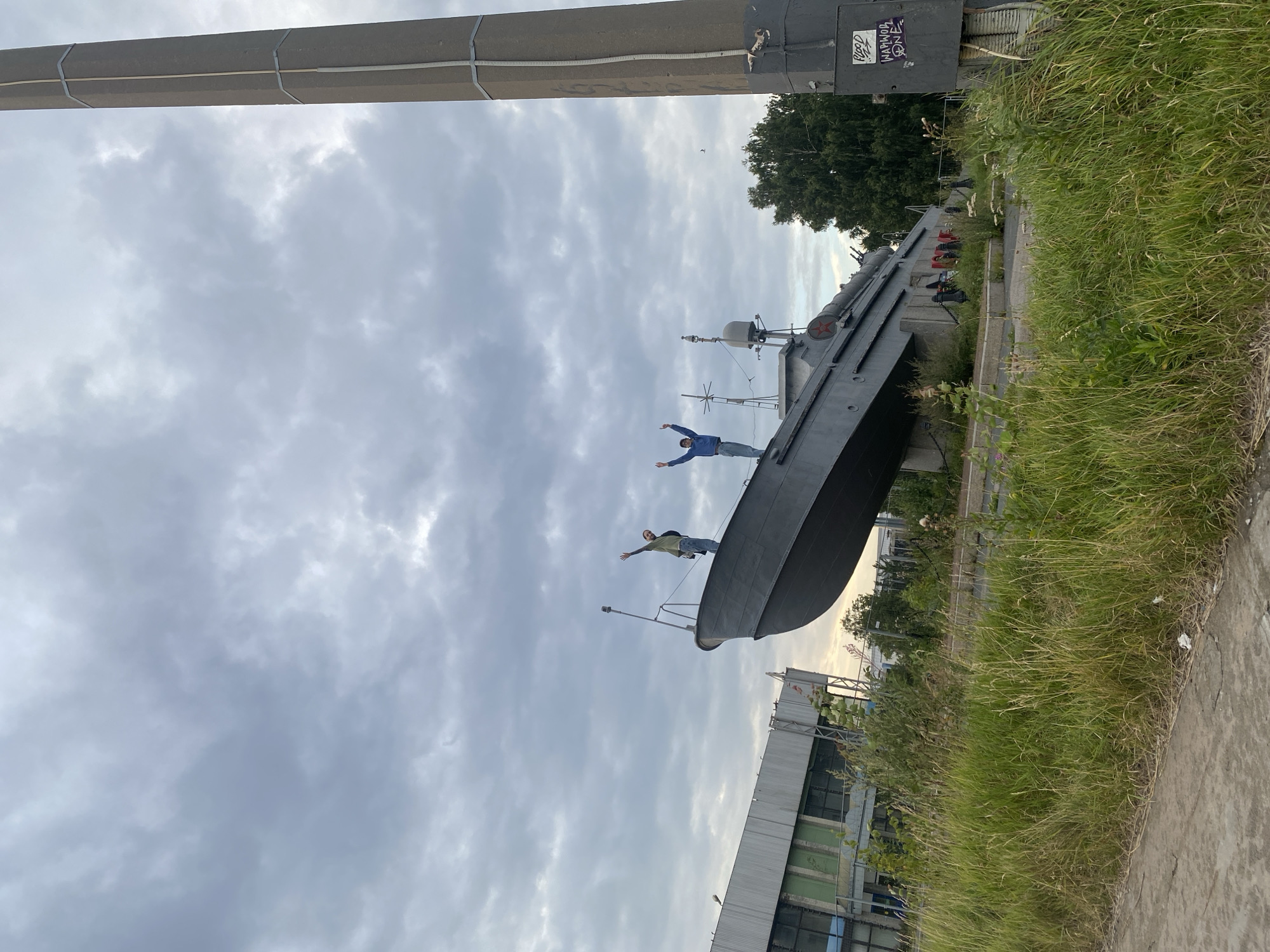 An outdoor, low-angle shot shows a gray ship, seemingly a former naval vessel, on display at a memorial site. Two individuals are standing on the deck of the ship, both with their arms raised in the air. The ship is situated on land, with tall grass and weeds growing around its base. In the background, a large, industrial-looking building with a silver facade and some green accents is visible on the left. To the right of the ship, a tall, cylindrical concrete pillar stands prominently. The sky is overcast with heavy gray clouds, suggesting a possibly moody or stormy atmosphere. The overall impression is one of a somewhat neglected, yet historically significant, outdoor monument. The location is likely Saint Petersburg, Russia, known for its rich maritime history and numerous memorials to naval achievements.