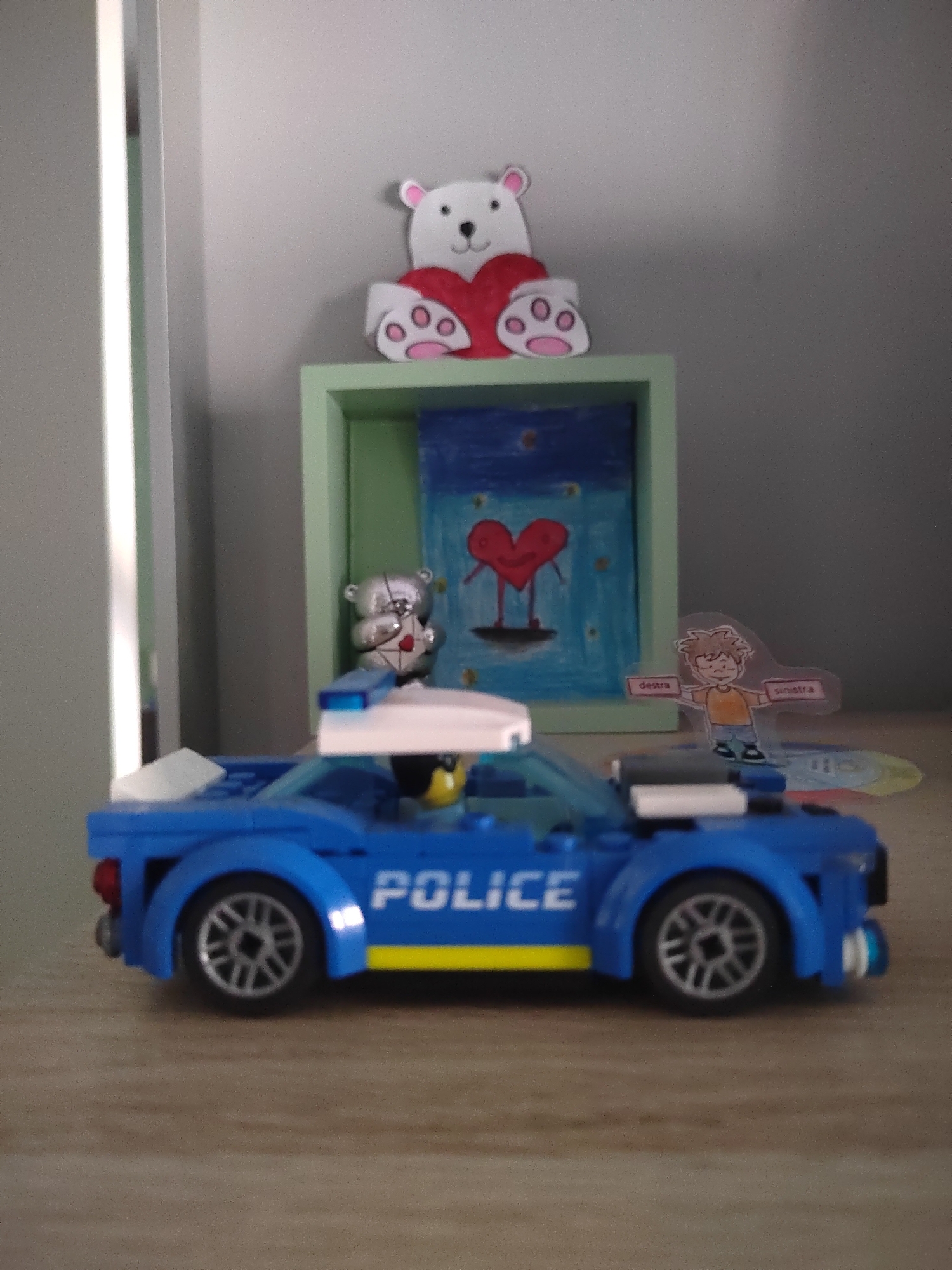 The image shows a shelf with various decorative items. On top of a green shelf, there is a white teddy bear with a red heart. Below it, inside the shelf, is a framed drawing of a red heart with legs. To the right of the shelf, there is a cutout of a boy with text that appears to say "destra" and "sinistra" on pink banners. In front of the shelf, a blue Lego police car is positioned. The Lego car has "POLICE" written on its side and a yellow stripe. The background appears to be a wall. There is no discernible information in the image that would indicate its location in Porto Recanati, Italy, or any other specific city.