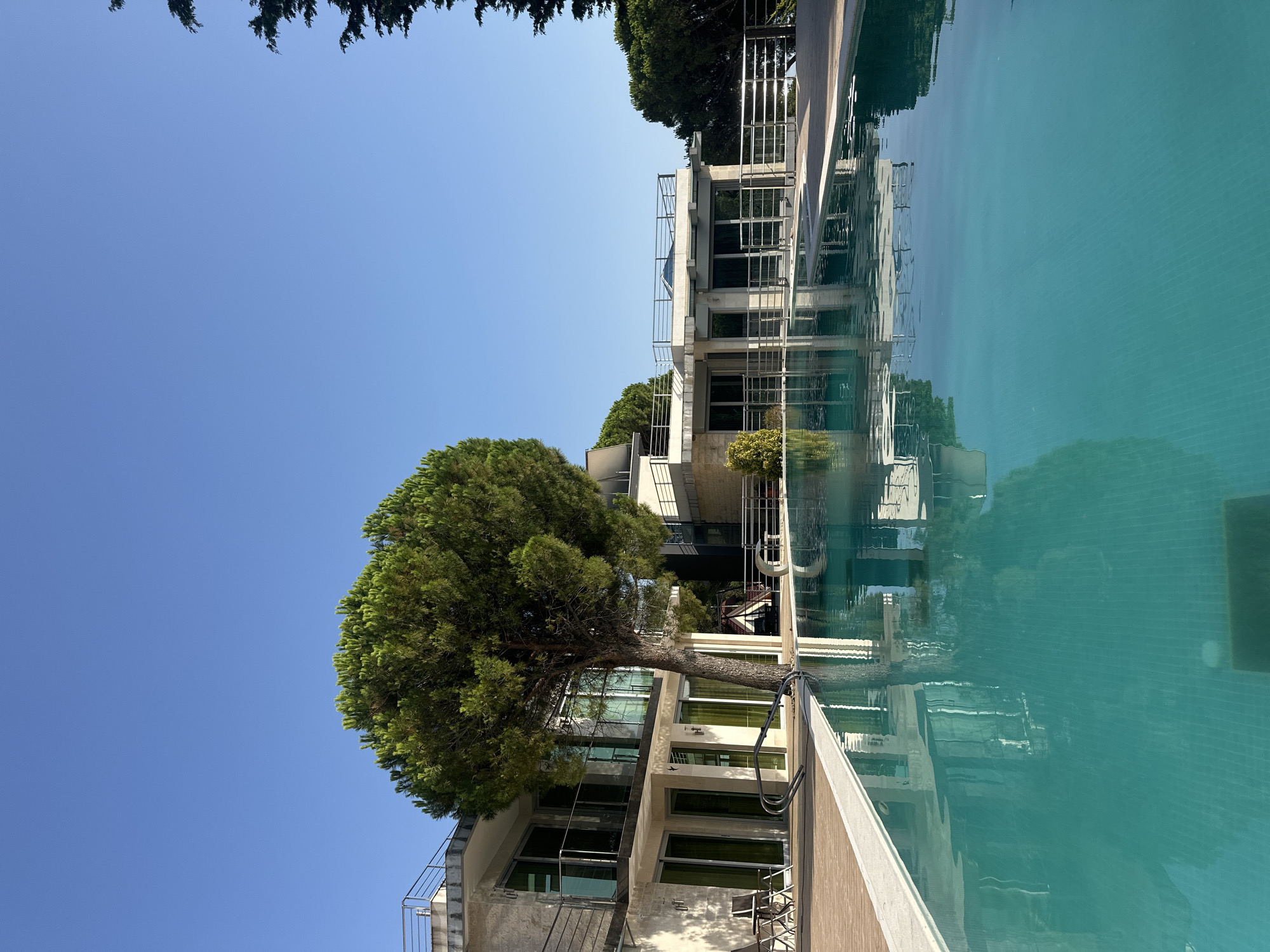 The image depicts a bright, sunny day in Brca, Montenegro. In the foreground, a large, turquoise swimming pool with tiled bottom reflects the surrounding environment. The pool is bordered by a light-colored deck made of wood planks.
To the left of the pool, a modern building with multiple levels is visible. The building features light-colored stone walls, large glass windows, and balconies with metal railings. A mature pine tree stands prominently in front of this building. Two outdoor chairs and a small table are placed on the deck near the pool.
Further back, another section of the building complex is visible, also with balconies and glass windows, set against a backdrop of lush green trees and a clear blue sky. The overall scene conveys a sense of luxury and relaxation, typical of a resort or private villa. There are no people or animals visible in the image.