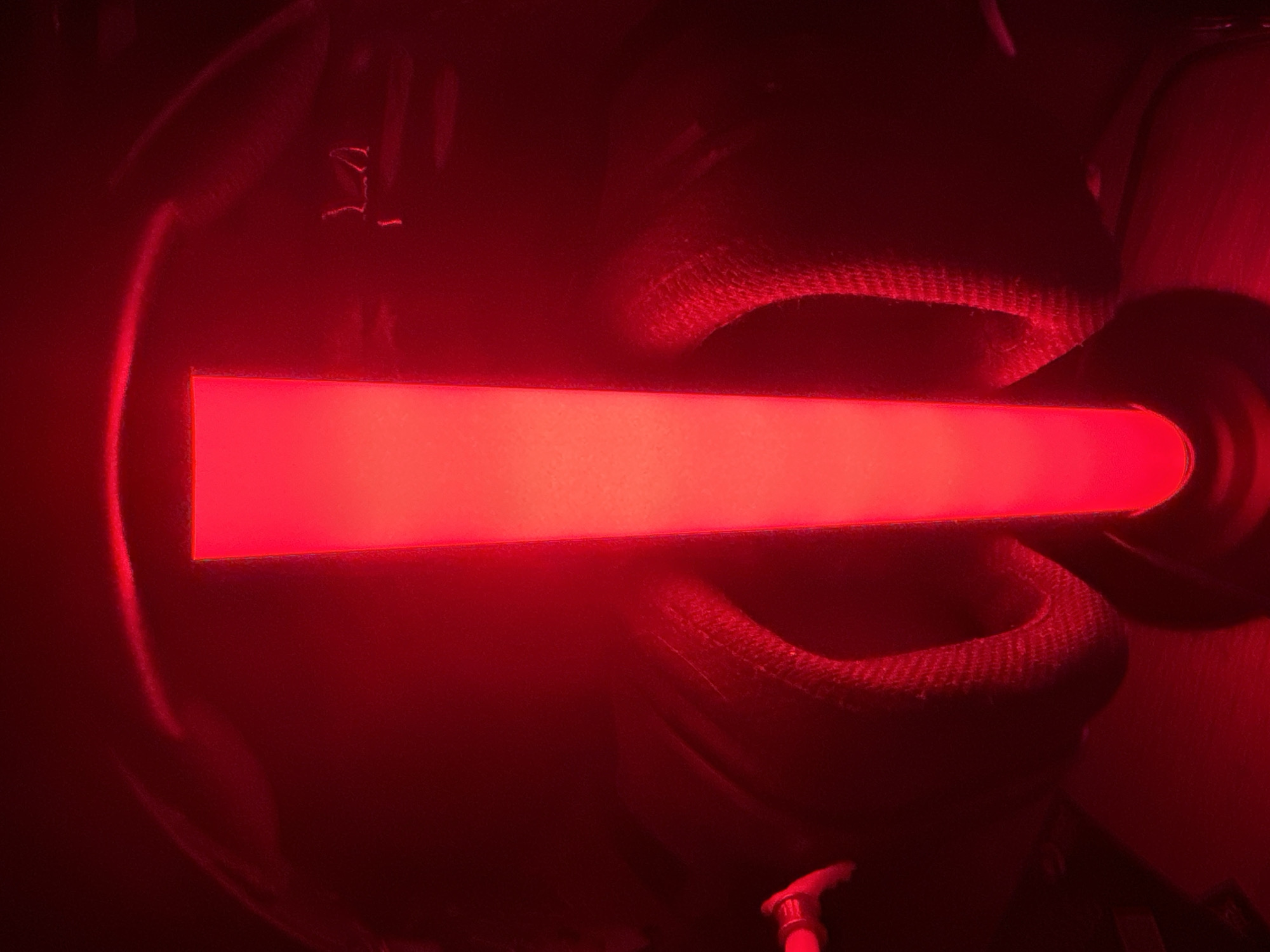 A bright red, illuminated rectangular object stands vertically in the center of a dark room. The object is surrounded by dimly lit, dark objects with textured surfaces, possibly fabric, that curve around its base. The lighting creates strong shadows and highlights, giving the scene an abstract and intense appearance. The overall impression is one of a close-up, dramatic shot focused on the glowing object.