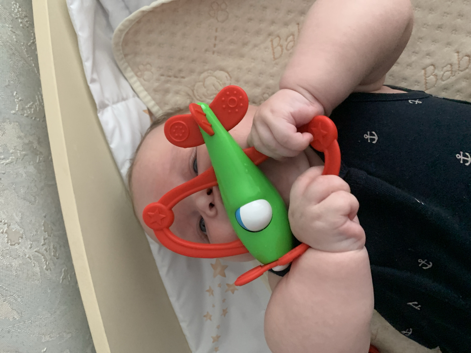 A close-up, overhead shot shows a baby lying on their back, holding a bright green toy airplane with red accents. The baby has pale skin and is wearing a dark blue garment with small white anchor patterns. The toy airplane has red propeller blades and a red handle. The baby's hands are gripping the toy, and their face is partially visible, with blue eyes looking towards the toy. The background consists of a white and patterned surface, likely a crib or bedding, with a textured wallpaper visible at the top. The lighting is soft, suggesting an indoor environment.