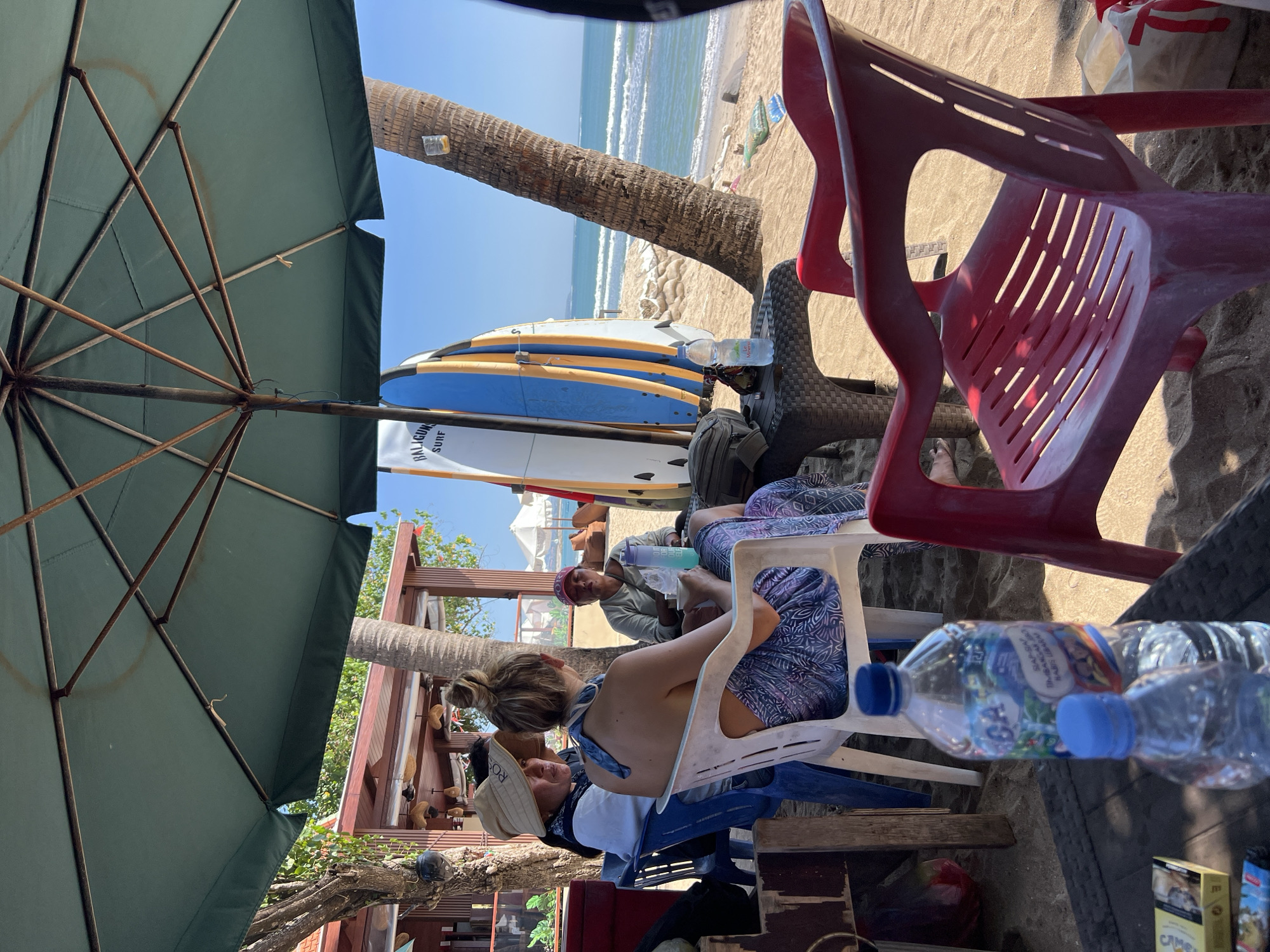 Underneath a large green beach umbrella, four individuals are seated on chairs on a sandy beach in Kuta, Indonesia. A woman wearing a wide-brimmed hat and a blue bandana is seated in a blue plastic chair on the left. Next to her, a woman with her back to the camera, wearing a patterned skirt and a blue bandana around her neck, is seated in a white plastic chair. To her right, a person in a tan hat and a light-colored shirt is seated in a blue chair, holding a blue cup with a straw. In the background, a portion of a white and blue surfboard with "BALIGUN SURF" written on it is visible. A small black table with a plastic water bottle and other items is located between the seated individuals. Beyond the beach, the ocean with gentle waves and a clear blue sky are visible. Two palm trees are present in the scene, one on the right side of the frame and another to the left, near a building with a red roof. A red plastic chair is in the foreground on the right.