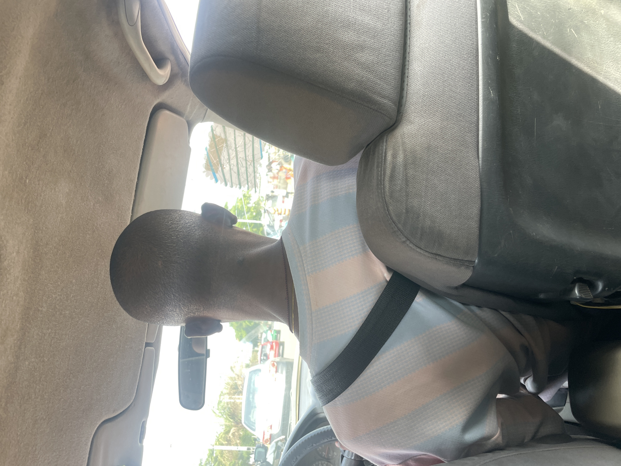 The image shows a person sitting in the driver's seat of a car in Abuja, Nigeria. The person has short hair, a gold necklace, and is wearing a striped shirt and a seatbelt. The car's interior is visible, including the dashboard, steering wheel, rearview mirror, and seat. Outside the car, other vehicles and buildings can be seen. The location is suggested to be Abuja based on the background.