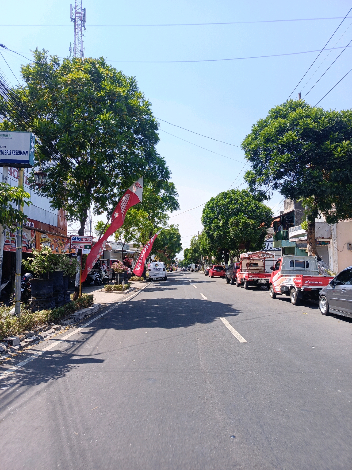 The image depicts a street scene in Magetan, Indonesia, on a clear, sunny day. The perspective is from the side of the road, looking down the street. On the left side of the road, there is a building with signage indicating "BPJS KESEHATAN" and "Alfamart" with a directional arrow indicating "200 M". Red flags with the Honda logo are visible, one prominently displayed in front of the Alfamart. Trees line the street, providing shade. A variety of vehicles are present, including several red pickup trucks, one of which has "HONDA" written on its side. A silver car is parked on the right side of the road. The road itself is paved with asphalt and has white lane markings. There are power lines and a cell tower visible in the background. The overall impression is of a busy commercial street in a town or city.