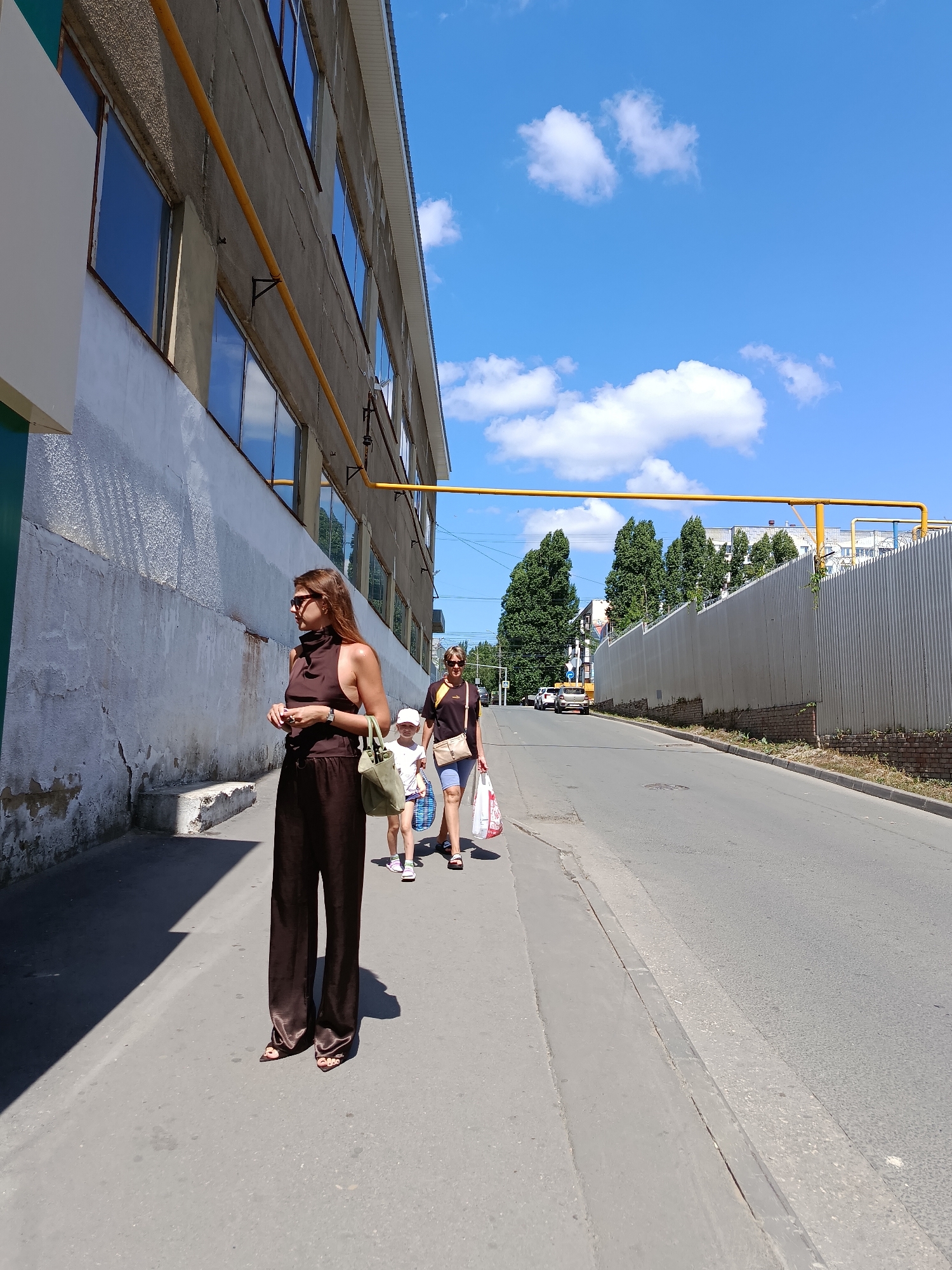 The image depicts a street scene in Saratov, Russia, on a sunny day. In the foreground, a woman with long brown hair wearing sunglasses and a dark brown sleeveless top with wide-leg pants walks on a sidewalk. She carries a dark green tote bag over her shoulder. Behind her, another woman and a young child are walking. The second woman wears a dark t-shirt and blue shorts, carrying shopping bags. The child, wearing a white cap and a light-colored t-shirt, walks alongside.
The environment features a large, industrial-style building with many windows on the left side of the image, characterized by a rough, light-colored textured facade and a visible yellow pipe running along its exterior. The sidewalk is paved and a low concrete curb separates it from the asphalt road. The road recedes into the distance, lined by a tall, corrugated metal fence topped with barbed wire on the right. Beyond the fence, several tall trees are visible, along with more distant buildings. The sky is bright blue with scattered white clouds. Several cars are seen on the road further in the distance. The overall impression is that of an urban industrial area on a clear day.