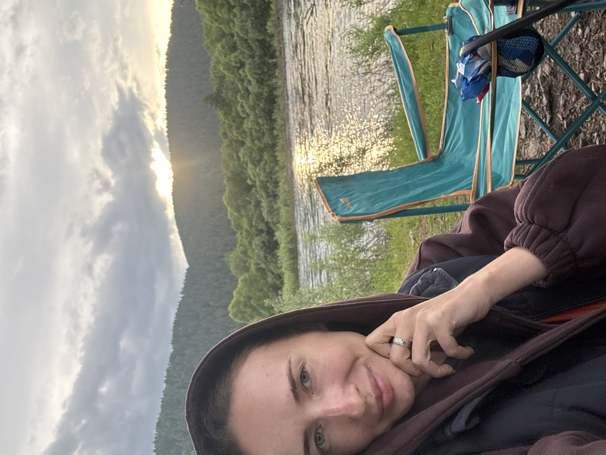 The image shows a woman posing for a selfie outdoors, with a river and forested hills in the background. The woman is wearing a hooded sweatshirt and a vest, and is resting her chin on her hand, which is adorned with a ring. To her right, a teal camping chair is visible, with some items in its side pocket. The sun is setting behind the hills, casting a warm glow on the water. The location is not specified as being in the city of Narva, Russia; rather, it depicts a natural outdoor setting.