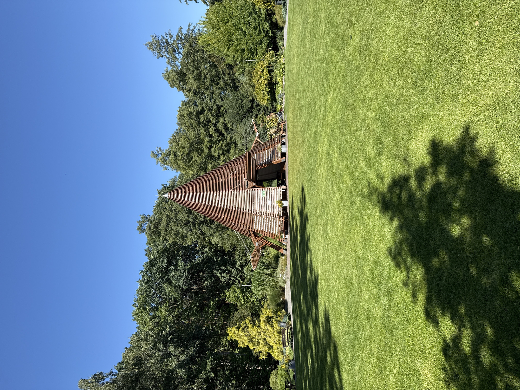 This is a photograph of the "Pod Zielonym Stojakiem" (Under the Green Stand) pavilion, located in the Botanical Garden of the University of Zielona Góra in Poland. The image captures the unique, conical wooden structure with its slatted design, which appears open on one side, revealing an interior space. The pavilion is situated on a vast, meticulously maintained green lawn. In the background, a dense forest of lush green trees forms a natural backdrop under a clear blue sky. To the left of the pavilion, a bright yellow ornamental tree stands out, accompanied by a park bench and some smaller plants. The foreground is dominated by the vibrant green grass, with dappled shadows from overhead foliage cast across its surface. The scene is peaceful and showcases a blend of natural and architectural elements.