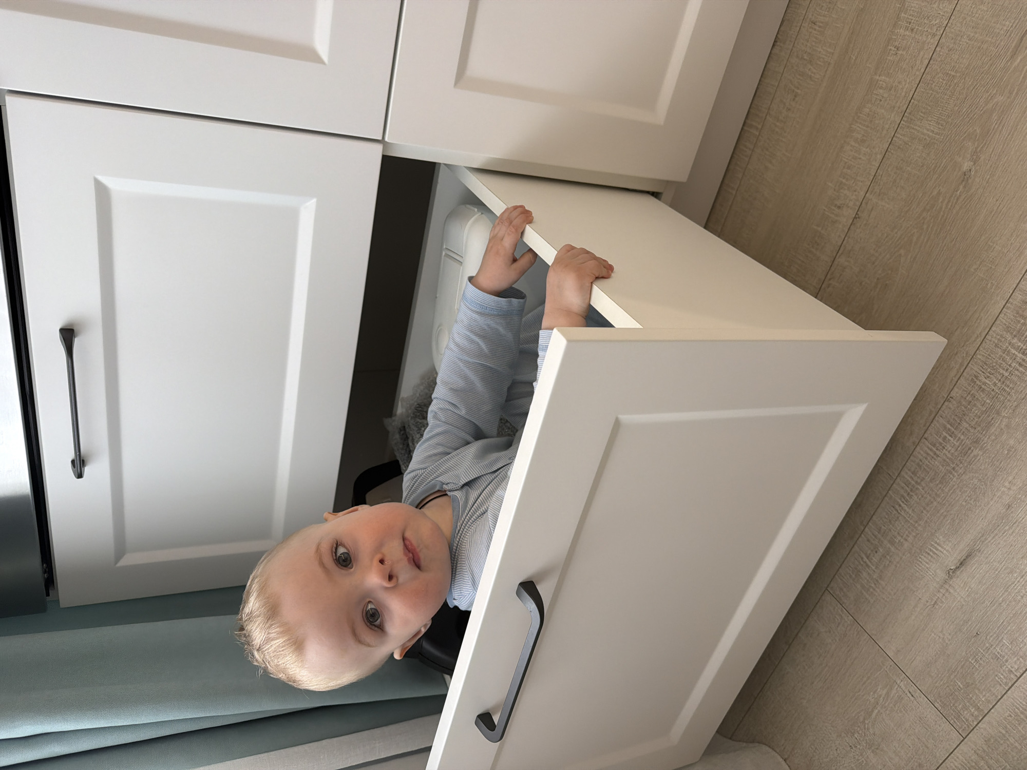 A toddler is partially inside an open white cabinet drawer. The drawer is open, and the child is visible from the chest up. The child has blonde hair and is wearing a light blue and white striped shirt. The cabinet is white with black handles, and the floor is light-colored wood. There is a blue curtain visible on the left side of the frame. The location is not identifiable as Votkinsk, Russia.