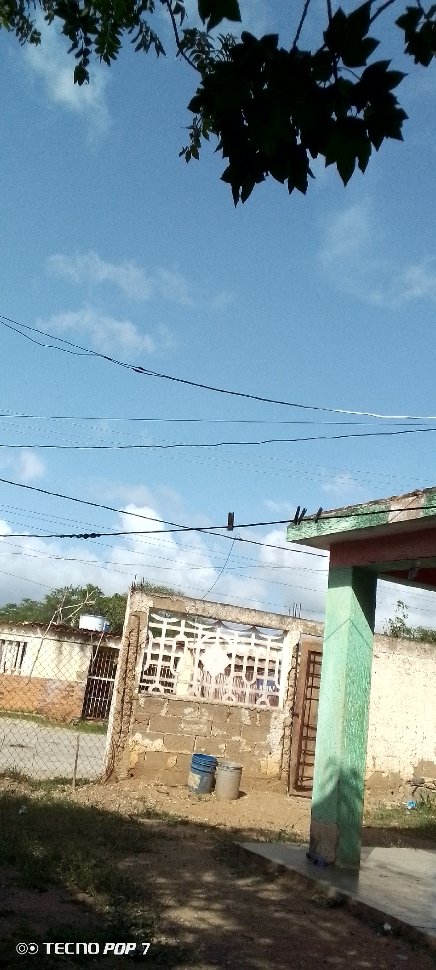 The image depicts an outdoor scene with a clear blue sky and scattered clouds. In the foreground, a section of a light-colored building with a green painted pillar is visible. A mesh fence runs along the front of a property. Beyond the fence, there is a building with a white, decorative screen wall and another section of a building with a metal-barred gate and a water tank on the roof. Two blue and gray plastic barrels are situated on the ground near the screen wall. Overhead, multiple power lines are strung across the sky. Dark green foliage from a tree hangs down from the top of the image. The text "TECNO POP 7" is visible in the bottom left corner. The scene appears to be in Altagracia, Venezuela, suggested by the phone branding which is common in that region.