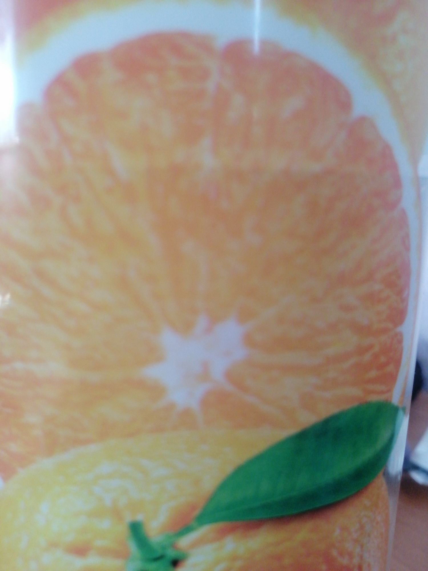 The image shows a close-up of a sliced orange, with a green leaf in the foreground. The orange is the dominant subject, filling most of the frame. Its segments and pith are clearly visible, with a bright, juicy appearance. The green leaf is positioned in the lower right quadrant of the image, resting on the orange. The background is blurred and appears to be a light color, possibly a white surface. The lighting is bright, highlighting the texture and color of the orange. The image is abstract and focuses on the details of the fruit. It is not possible to determine the location in Milzkalne, Latvia from the image itself.