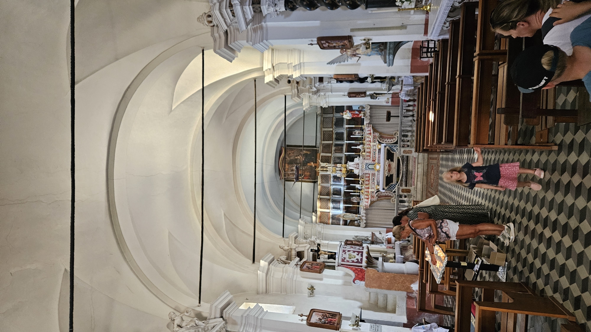 The image shows the interior of a church, likely in Sant'Antonino, France, with an ornate altar at the far end. Several people are present. In the foreground, a young girl with blonde hair, wearing a dark blue t-shirt with a butterfly design and a pink skirt, stands near the aisle. To her right, an adult woman with blonde hair is visible from the back, wearing a dark baseball cap. In the mid-ground, two women are standing near a candle-lighting station; one is wearing a patterned top and white shorts, and the other is wearing a long, dark patterned dress. The church features wooden pews on either side of a checkered floor. The architecture includes arched ceilings and decorative elements on the walls and columns.