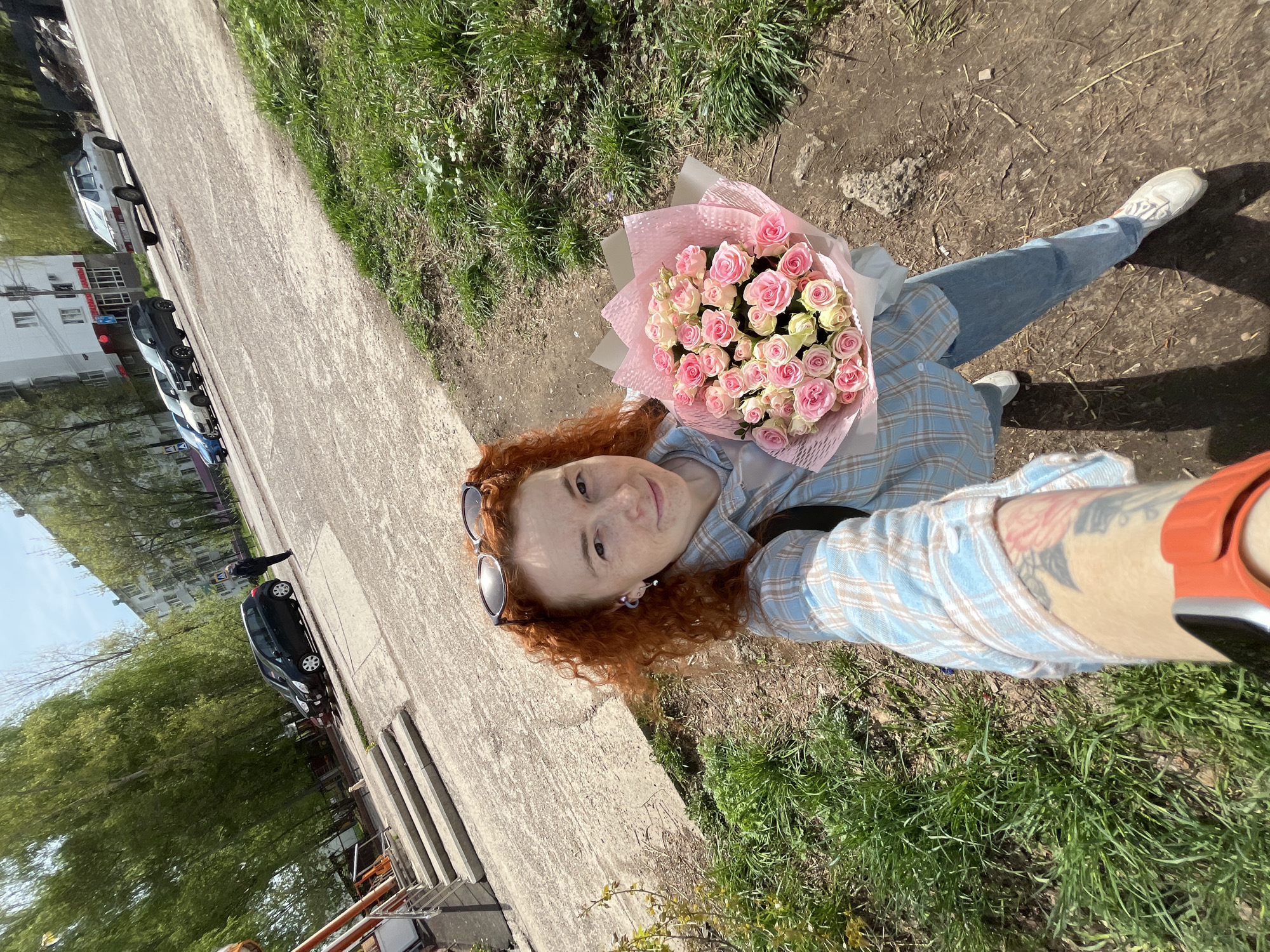 A fair-skinned woman with curly red hair takes a selfie. She holds a bouquet of pink and white roses wrapped in pink paper. She wears a light blue and tan plaid shirt, blue jeans, and white sneakers. Sunglasses are perched on her head. Part of a tattoo is visible on her forearm.
The woman stands on a sidewalk next to a grassy area. A street with parked cars and buildings is visible in the background. The location is Chaikovsky, Russia.