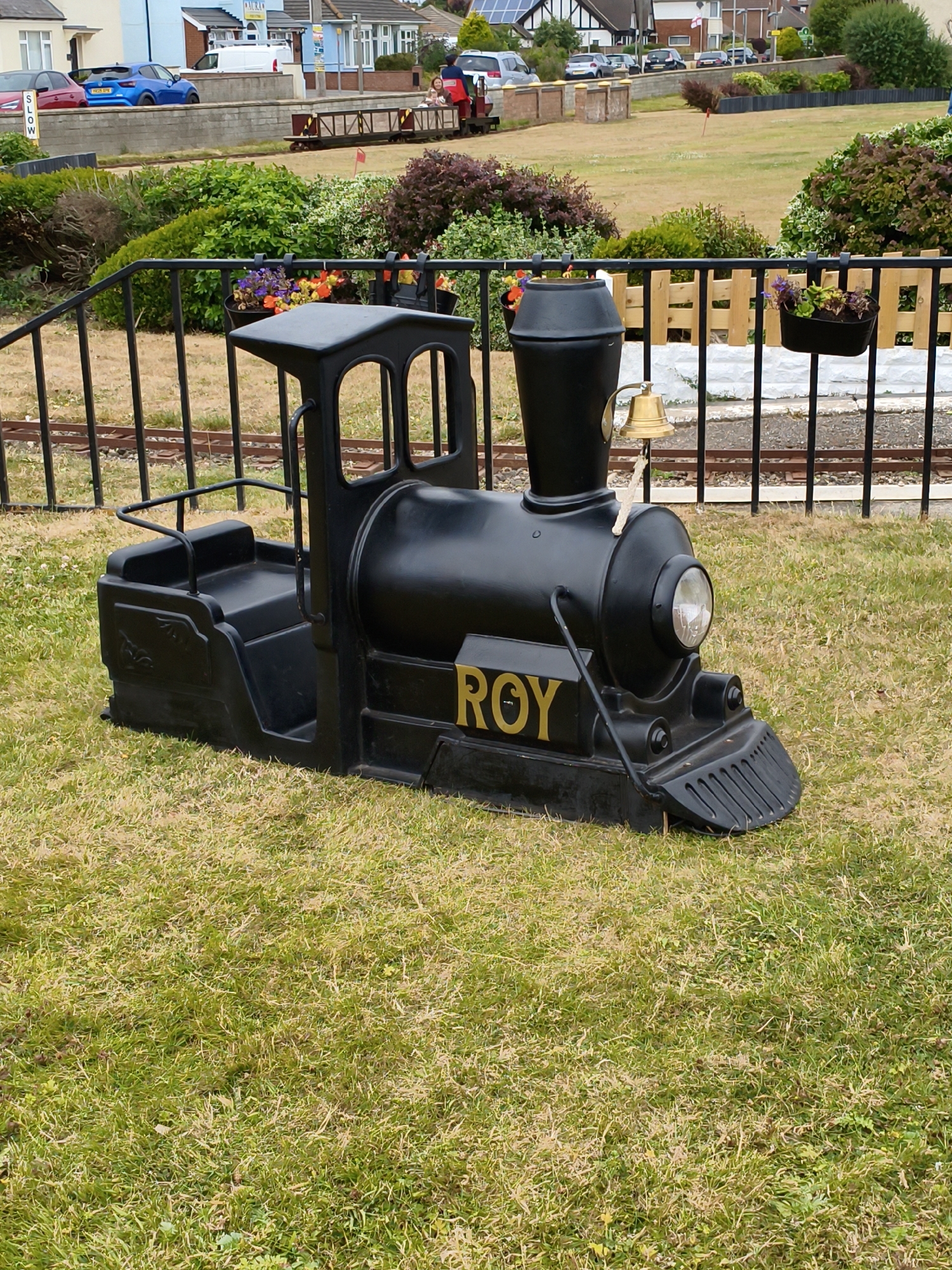 The image shows a black toy train labeled "ROY" sitting on a grassy area. In the background, there is a miniature railway track, a fence, and some residential buildings. There are also some people visible on the miniature train in the background. The location is identified as East Lindsey, United Kingdom. The toy train has a bell on top and a headlight. The grassy area appears to be dry in some spots, indicating it might be summer. The overall scene suggests a recreational park or a place with a miniature railway attraction.