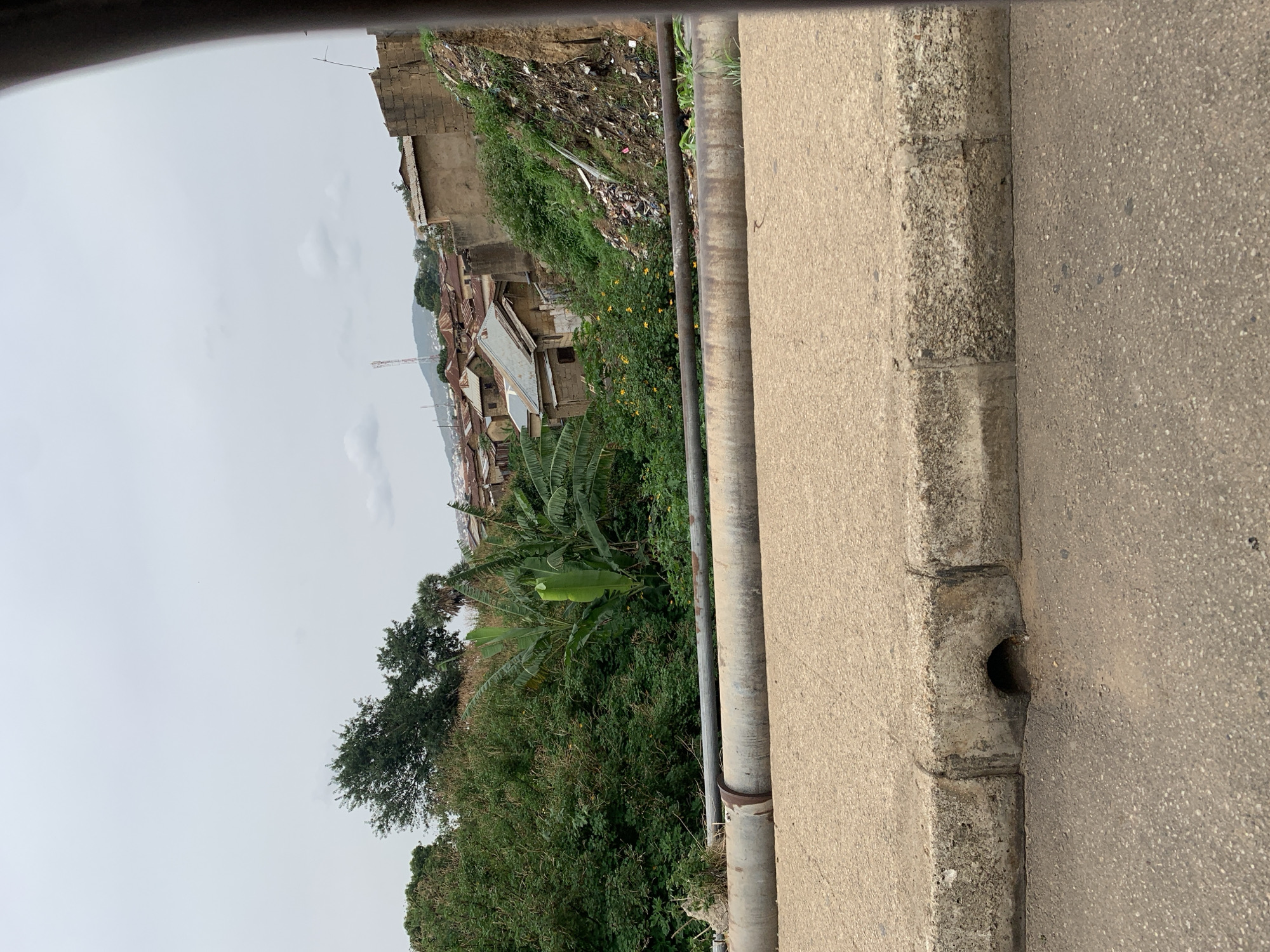 View from a roadside in Kabong, Nigeria. A concrete curb separates the paved road from a steep slope covered in lush green vegetation and some buildings. In the background, rooftops of houses are visible on a hill. The sky is overcast. A drain opening is located in the curb. Two metal pipes run along the curb. The hillside contains patches of trash.
