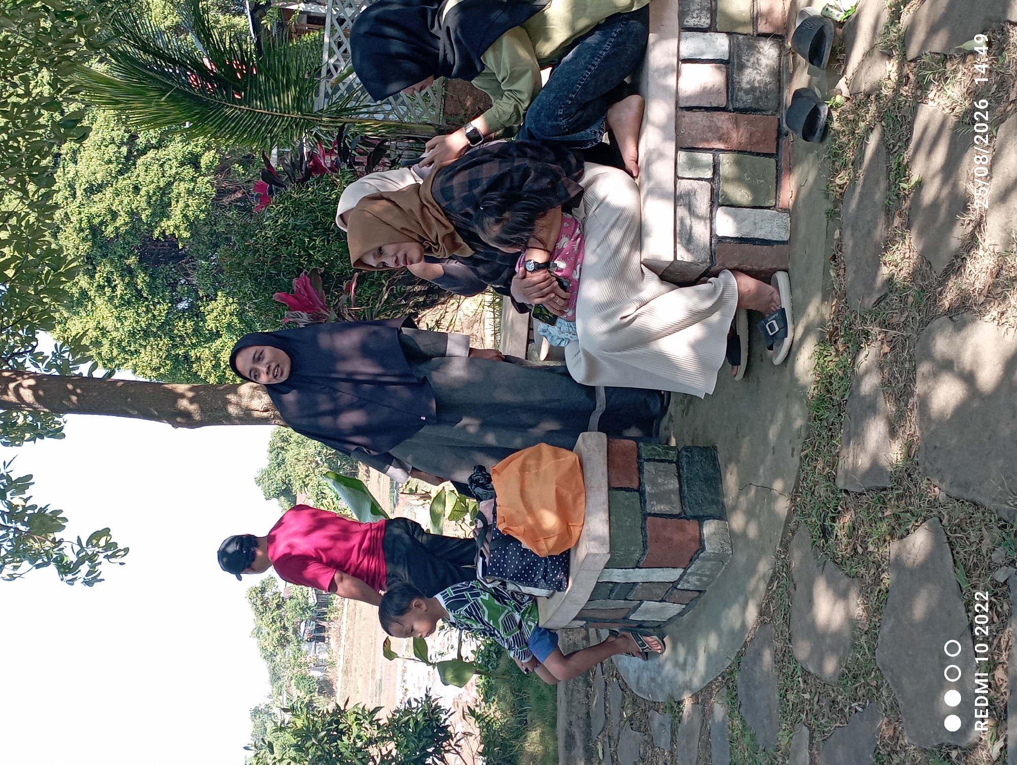 In a park setting in Tayu, Indonesia, on a bright, sunny day, a group of people are gathered. A man in a red shirt and black cap stands in the background, looking towards the left. To his right, a woman in a black hijab and grey dress is speaking and gesturing. In the foreground, a young boy in a green and black striped shirt and blue shorts sits on a stone bench, looking to his left. Beside him are two bags, one orange and one patterned. Further to the right, on the same bench, three women are seated. One woman in a brown hijab is holding a baby. Another woman in a beige hijab is seated next to her, and a third woman in a black hijab is leaning on the first woman's shoulder, appearing to speak to her. The bench is made of colorful stones. The ground is paved with large, irregular stones and patches of grass. Lush green trees and foliage surround the area, and a palm tree is visible on the right. The overall atmosphere is relaxed and social.