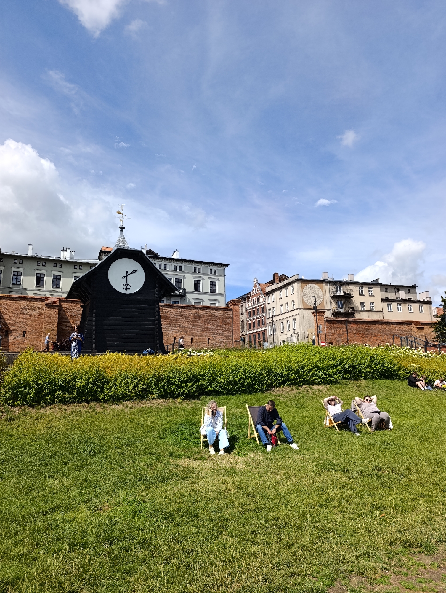 In the city of Toruń, Poland, a sunny day unfolds with people relaxing on a grassy hill. In the background, a historical clock tower with a weather vane stands tall against a blue sky with scattered clouds. Adjacent to the tower, a red brick wall encloses buildings with traditional European architecture. Green bushes and trees line the perimeter of the hill, adding a touch of nature to the urban landscape. Several individuals are seen lounging in deck chairs, enjoying the pleasant weather. One person is seen in a white shirt and blue jeans, another in a dark shirt and jeans, and a third in a light-colored shirt and dark pants. The overall scene depicts a peaceful and leisurely atmosphere in a picturesque city setting.