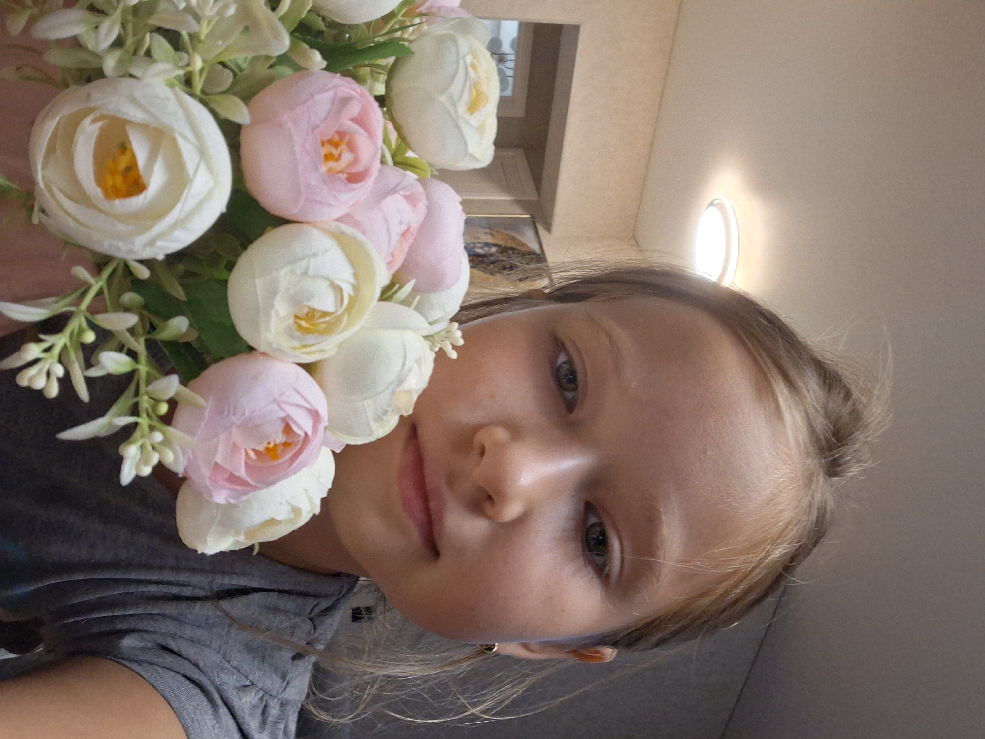 A young girl with blonde hair and brown eyes is holding a bouquet of artificial flowers. The flowers are a mix of white and pink, with green leaves. The girl is wearing a grey t-shirt. She is looking towards the camera, and her expression is neutral. The background is blurry, but it appears to be an indoor setting with a light-colored wall and a light fixture on the ceiling. There is also a glimpse of a framed picture on the wall in the background. The overall lighting suggests it is daytime, possibly indoors near a window or under artificial light. There is no visible text in the image. The photo appears to be a selfie, given the angle and the close-up nature of the shot. The girl's pose and the way she is holding the flowers suggest she might be posing for the photo.