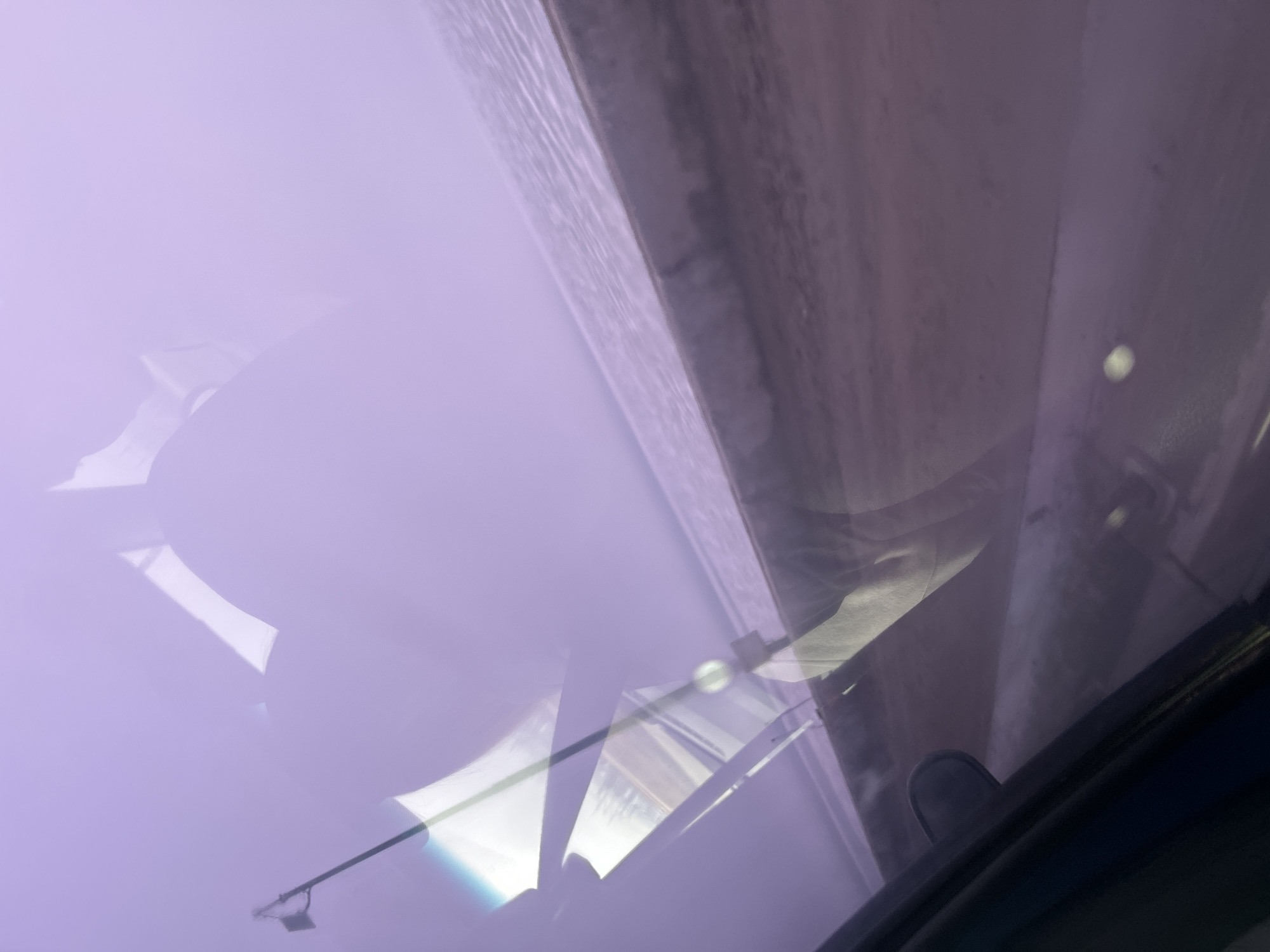 The image appears to be taken from inside a car looking upwards through the sunroof/window. Visible through the glass are reflections and distorted views of the surrounding environment. A portion of a building and window structures are seen on the right side of the frame, with a blurred reflection of what appears to be a traffic light or other light source. The color cast is purplish. No people are visible. It's impossible to confirm the location within Chaikovsky, Russia based on the available visual information.