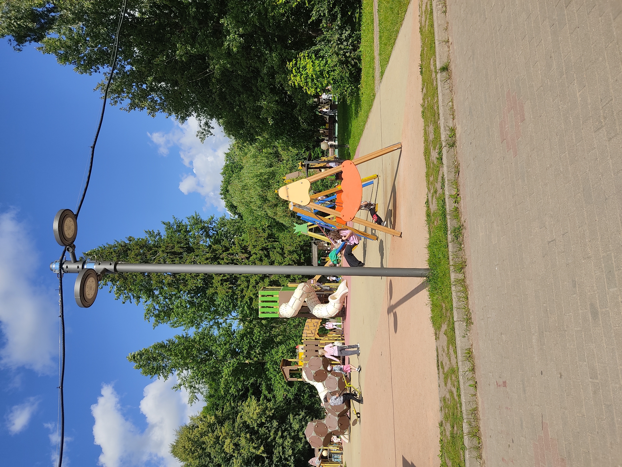 The image depicts a playground in a park on a sunny day, with a clear blue sky and scattered white clouds. In the foreground, a paved walkway runs horizontally, bordered by a strip of grass and a curb. Behind the walkway, a sandy-colored playground area is visible, featuring various play structures including slides, swings, and climbing equipment. Several children are present, engaged in play. A lamppost with two modern LED lights stands prominently on the left side of the playground. The background consists of lush green trees, suggesting a summer or early autumn setting. The overall scene is one of a vibrant and active public space within a city park.