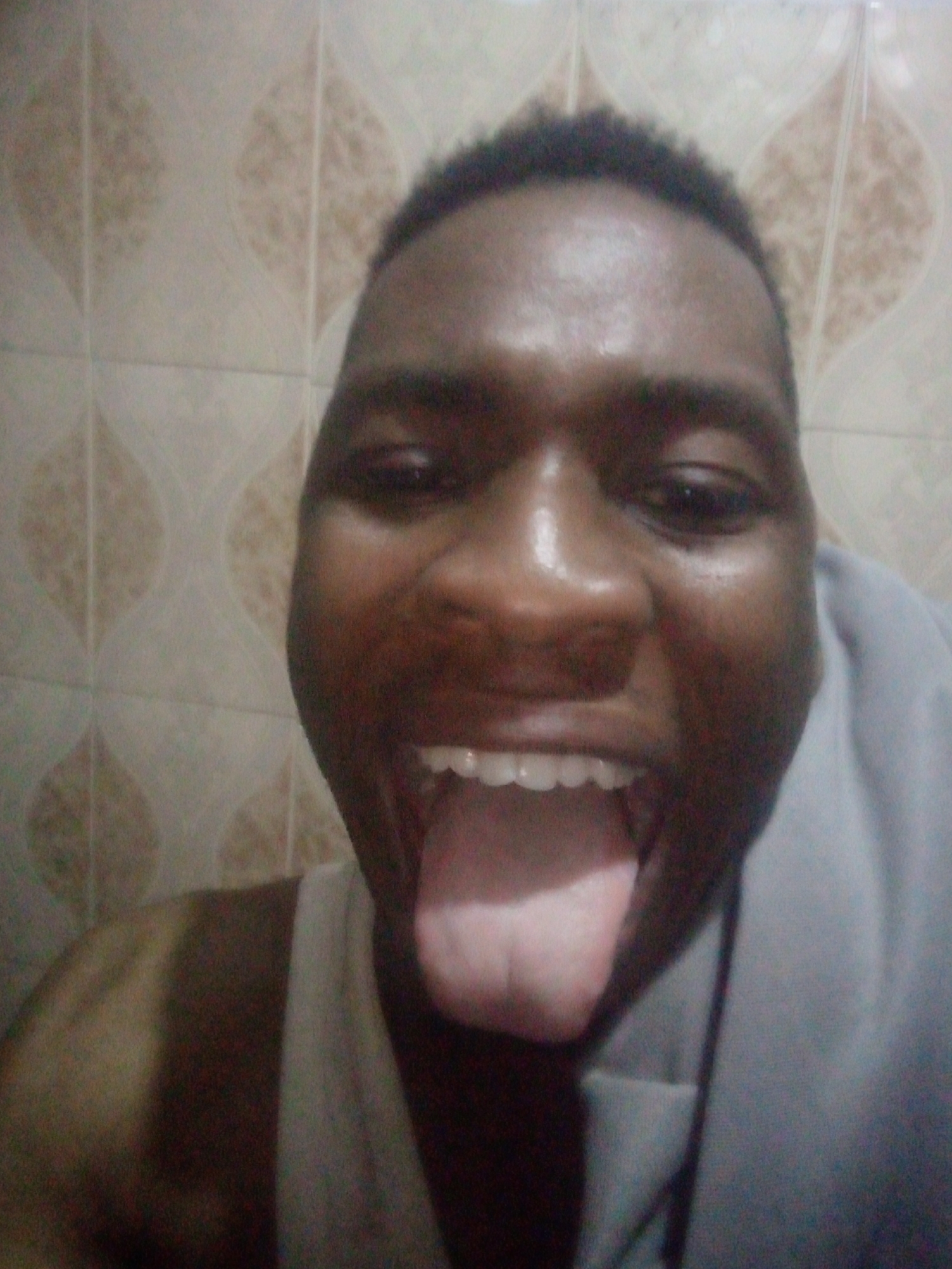 A close-up shot depicts an individual with dark skin, short, dark hair, and exposed teeth and tongue. The person is wearing a white tank top, and part of a grey shirt or cloth is visible on the right side of the frame. The background consists of tiles with a repeating leaf-like pattern. The location is Unknown, Unknown.