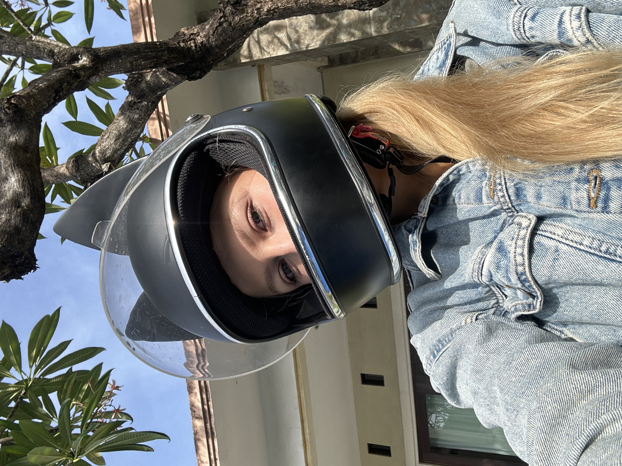 A woman wearing a black helmet with a transparent visor and a denim jacket is visible. She has long blonde hair. The background suggests an outdoor setting with a tree branch and leaves visible at the top left. A building with windows can be seen in the background. The location is Kuta Selatan, Indonesia.