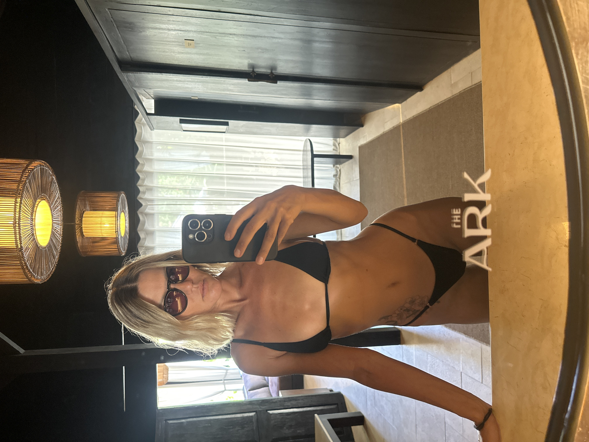 The image displays a mirror selfie of a female in a room in Mengwi, Indonesia. A woman with shoulder-length blonde hair and sunglasses stands centered, wearing a black string bikini consisting of a triangle top and a thong bottom. She holds a dark-colored smartphone with a triple-lens camera setup in her right hand, angled towards the mirror to capture her reflection. A tattoo, appearing to be floral or organic in design, is visible on her left hip/lower abdomen. Her left forearm rests on a light-colored, possibly marble or stone, vanity surface in the foreground.
The room, as reflected in the mirror, features a dark-colored ceiling from which two prominent circular pendant lights with a woven or rattan design emit a warm yellow glow; a third, smaller similar light is visible further back. Dark wooden doors or panels are present on both sides, suggesting built-in cabinetry or room dividers. Through an open doorway on the left, a dark sofa and another table are visible. White, translucent curtains cover a window or glass door in the background, allowing diffuse natural light to enter and hinting at greenery outside. The floor combines light-colored tiles and a dark-colored rug. In the lower center of the mirror's reflection, reversed white text reads "THE ARK" on the light-colored vanity surface.