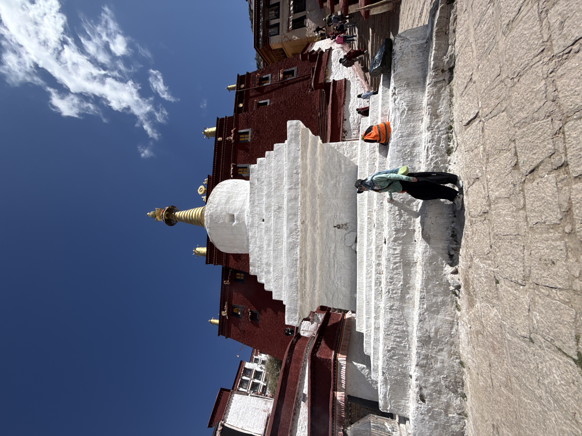 The image captures a scene at the Potala Palace in Lhasa, China. The architecture combines white and red facades and stepped structures. A central, white stupa with a golden spire is a focal point of the image, set against a clear blue sky with some white clouds.
Several people are visible in the image. One person, wearing dark trousers and a light-colored jacket, is climbing the white steps of the structure. Further up the steps, there are other individuals, some wearing brightly colored clothes.
The environment is defined by stone and concrete textures, suggesting a historical site frequented by tourists and locals alike. The overall impression is a sunny day at a significant cultural landmark.