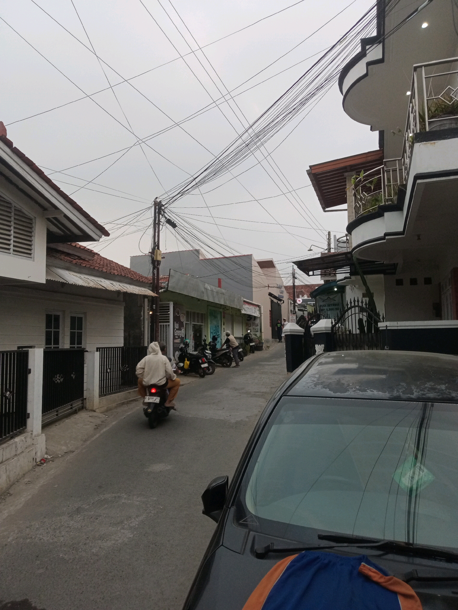 A slightly overcast day in Grendeng, Indonesia, is captured in this street-level photograph. The scene depicts a narrow asphalt road lined with residential buildings and small shops. A black car is partially visible in the foreground, its windshield reflecting the sky and overhead electrical wires.
The focus of the mid-ground is a person riding a motorcycle, wearing a light-colored hooded jacket. The motorcycle is moving away from the camera. Several other motorcycles are parked along the roadside, suggesting a communal living area or a place with local businesses. The architecture of the buildings shows a mix of traditional and modern elements, with tiled roofs and balconies. Electrical wires are a prominent feature, strung haphazardly across the sky and between poles, indicating a densely wired urban environment. The overall impression is of a typical Indonesian street scene, active with daily life and transportation.