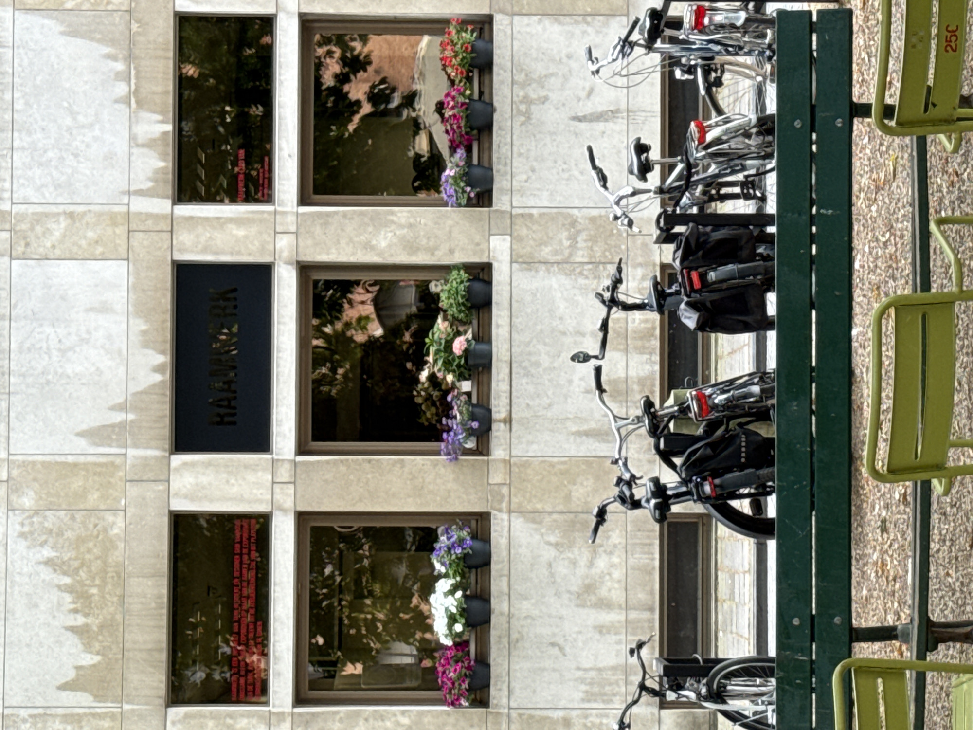 The image depicts the facade of a building with a stone exterior, divided into three horizontal sections. The top section features two large, dark-colored recessed areas with text. The left one displays red text, while the right one has "HAAMWERK" in white lettering. The middle section contains three square windows above planters filled with various flowering plants, including pink, white, and purple flowers. The bottom section of the building has a row of parked bicycles. In the foreground, a dark green bench is visible, with two light green chairs on either side, and an area of gravel on the ground. The scene appears to be outdoors in an urban environment. The building's architecture and the presence of the text "HAAMWERK" suggest a location in Bruges, Belgium, as this term is associated with a former industrial site in the city.