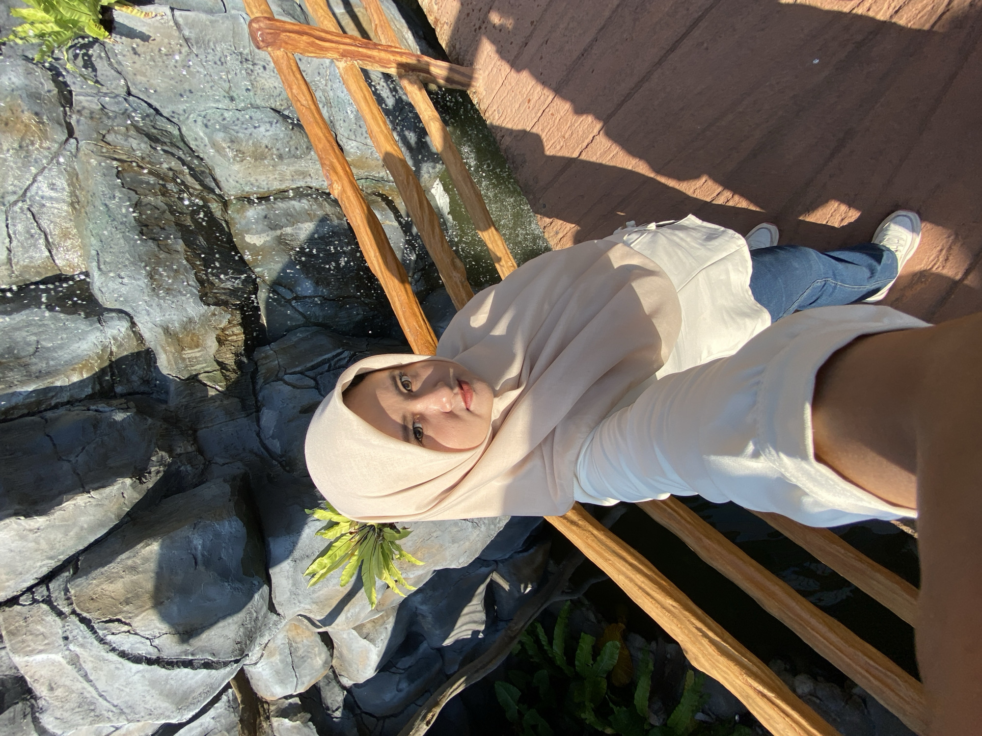 A person wearing a hijab, a white shirt, and blue jeans is taking a selfie on a bridge-like structure. The structure has wooden railings. Below the railings is a faux rock formation and water, indicating the location may be a water feature or artificial waterfall. Green plants are growing near the rocks. The background includes a brick-like surface and sunlight, suggesting an outdoor setting. The photograph does not indicate the location is in Mangli, Indonesia.