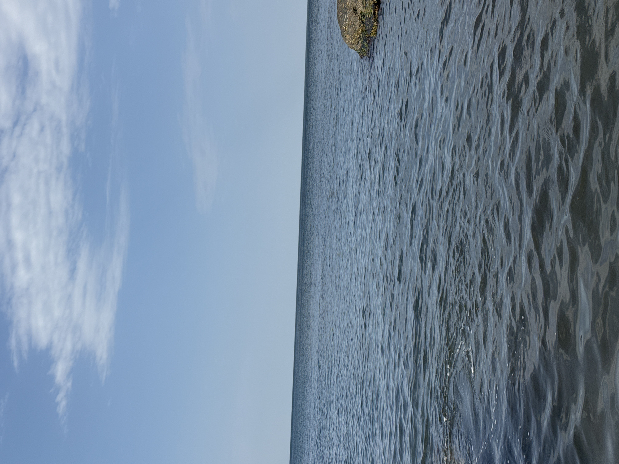 The image depicts a tranquil seascape under a partly cloudy sky. The horizon line is clearly visible, dividing the calm, dark blue water from the pale blue sky. A few scattered white clouds are present in the upper portion of the sky. On the right side of the image, a large, rough-textured rock, partially submerged in the water, is visible. The water exhibits gentle ripples, suggesting a calm weather condition. The scene is devoid of any human presence or activity. This view is characteristic of the coastline of Aktau, Kazakhstan, a city situated on the Caspian Sea.