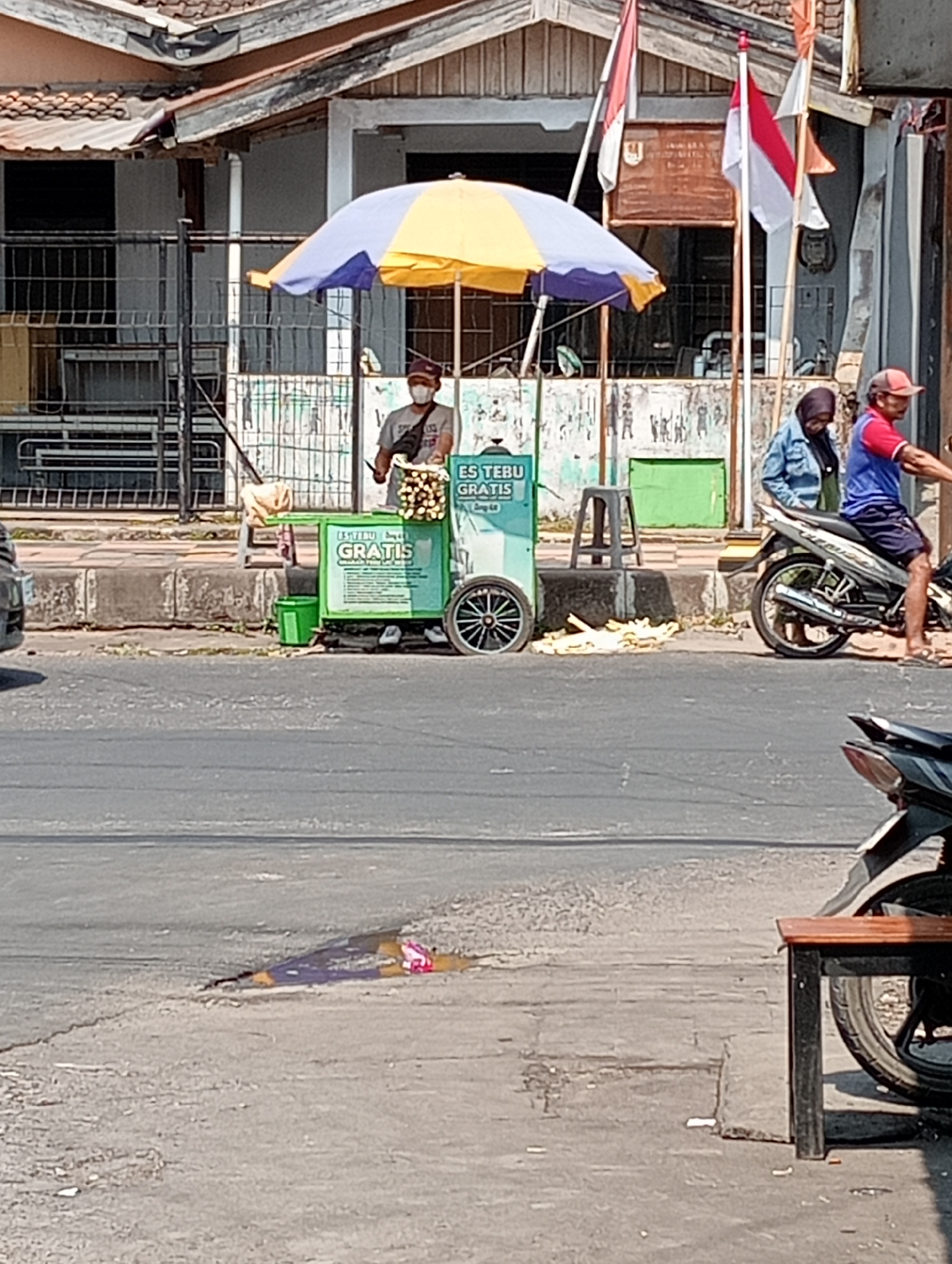On a sunny day in Grabag, Indonesia, a vendor stands beside a green ice cart, offering sugarcane juice. The cart is adorned with the words "Es Tebu Gratis," meaning "Free Sugarcane Juice." A yellow and purple umbrella shades the vendor and their cart. Several sugarcane stalks are neatly stacked on the cart, ready to be processed. In the background, a building with a tiled roof and several flags are visible, suggesting a public or commercial area. To the right, two people on a motorcycle are parked near the cart, possibly customers. The scene is set on a street with a sidewalk, and a road in the foreground. The overall atmosphere is one of a bustling street scene in Indonesia, with a focus on local commerce and everyday life.