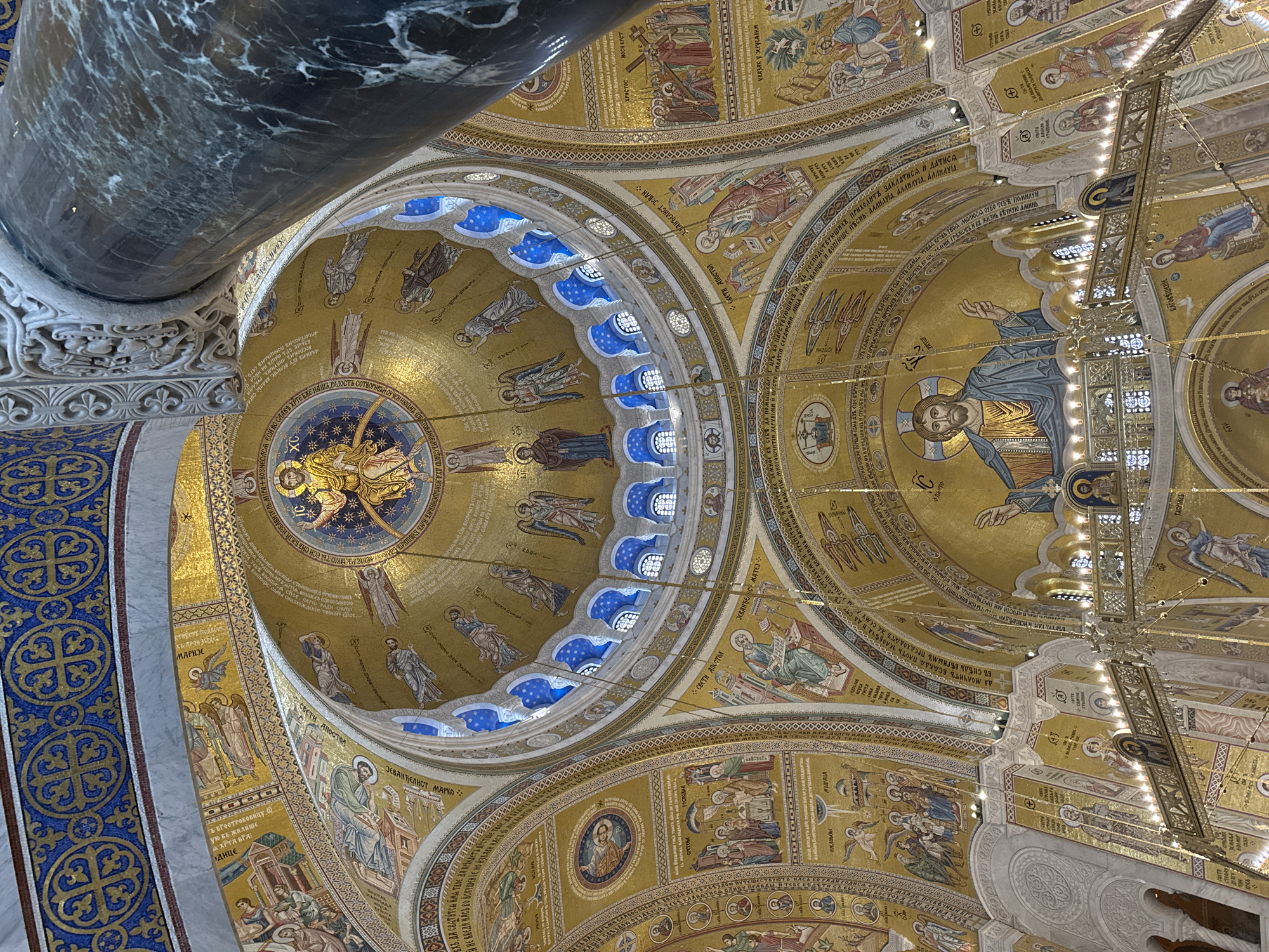 The image captures an interior view of the Church of Saint Sava in Belgrade, Serbia, focusing on the intricately decorated ceiling and domes. The architecture is Byzantine-inspired, with gold mosaic artwork covering the curved surfaces. The central dome features a representation of Christ Pantocrator surrounded by angels and biblical figures, with smaller surrounding domes depicting various saints and scenes from the New Testament. A large, ornate chandelier hangs partially in the frame, adding to the grandeur of the space. The image is taken from a low angle, looking upwards, emphasizing the height and scale of the church. A massive marble pillar with ornate carvings is visible on the left, framing the scene. The use of gold and vibrant colors, combined with the sheer scale of the artwork, creates a sense of awe and reverence. The image is a detailed depiction of the artistic and architectural elements within one of the most prominent religious buildings in Serbia.