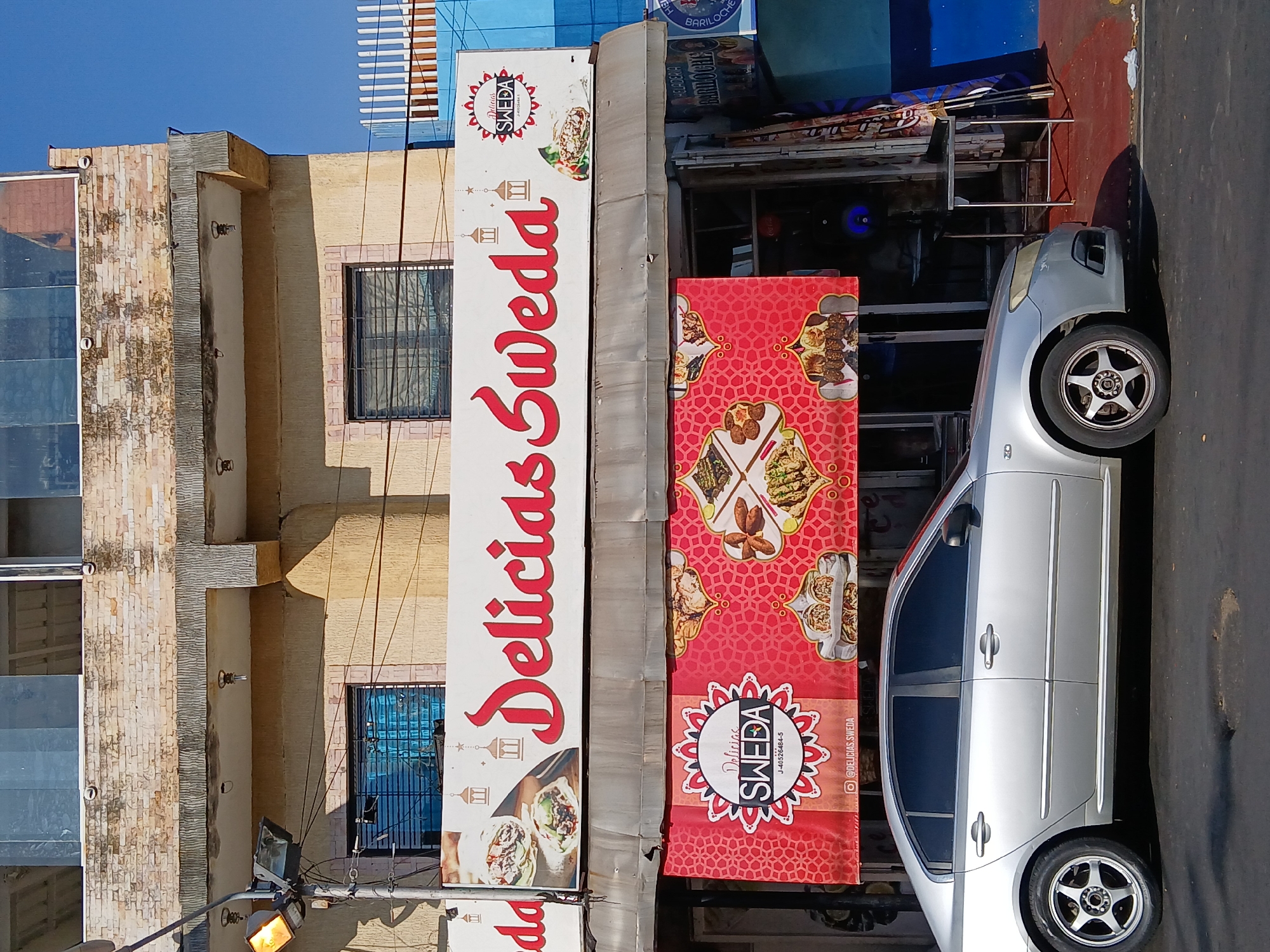 This image captures a bright, sunny street scene in Ciudad Guayana, Venezuela, likely a commercial district. The primary focus is a business named "Delicias Sweda," heavily advertised through prominent signage.
On the left, a multi-story building features a textured, light-colored facade, possibly brick or stone, with large reflective windows. Adjacent to it, another building, which houses "Delicias Sweda," has a similar light-colored facade and a window secured with dark metal bars.
A large, vertical white sign on the building prominently displays "Delicias Sweda" in bold red script, accompanied by images of food, likely shawarmas or wraps, and decorative lantern motifs. Next to this sign, a large red banner stands vertically, showcasing an array of prepared dishes, possibly Middle Eastern or Mediterranean cuisine. This banner also features a circular logo for "Delicias SWEDA," the identification number "J-40526464-5," and the Instagram handle "@DELICIAS.SWEDA."
A silver four-door sedan, possibly a Toyota, is parked on the asphalt street directly in front of the red banner. Its wheels feature distinctive five-spoke silver alloy rims. Behind the car, a smaller sign is partially visible, indicating "HELADERIA BARILOCHE" and "BARILOCHE," suggesting an ice cream parlor.
The time of day is clearly daytime, with strong sunlight casting shadows and indicating clear weather. Overhead, power lines are visible, connecting to poles with streetlights. No people are visible in the frame. The scene portrays a typical urban commercial environment.