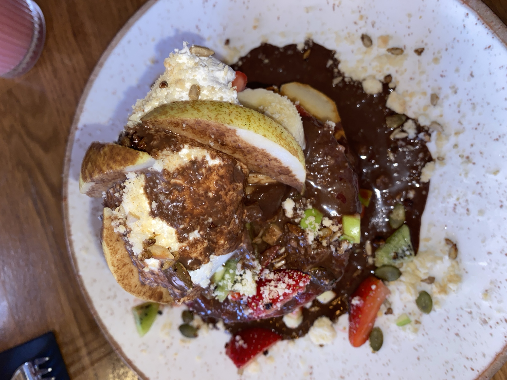 The image depicts a plate of dessert, likely served in Strasbourg, France. The dessert consists of a pancake topped with whipped cream, a pear, chocolate sauce, strawberries, kiwi, banana slices, and nuts/seeds. The plate is speckled with brown stains, and the background shows part of a wooden table. A blurred glass of pink liquid and a black object are also visible on the table.