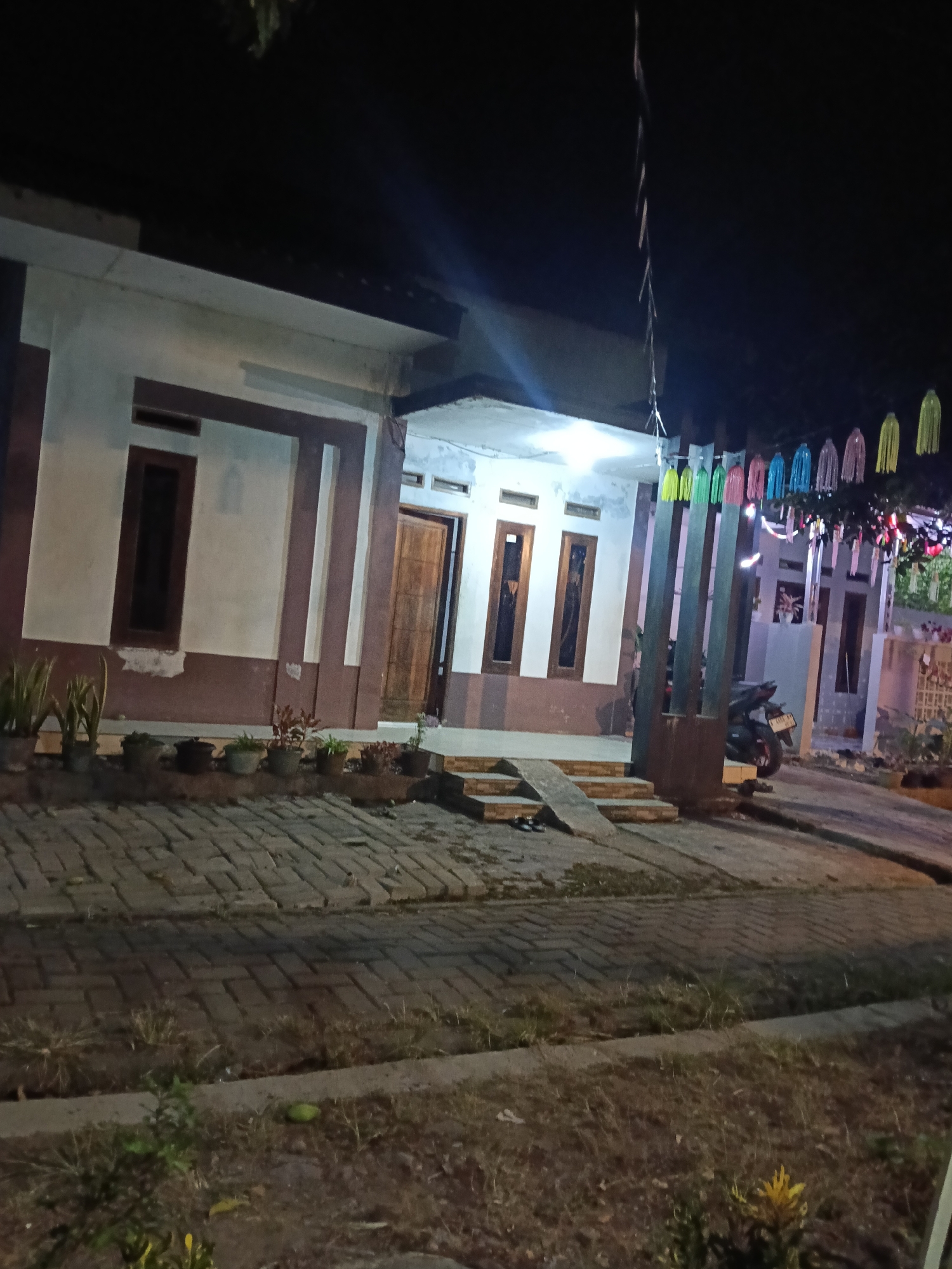 The image depicts the exterior of a house at night in Paojan Lor, Indonesia. The house has a light-colored facade with dark brown trim. There are three windows visible on the front of the house, two with dark frames and one with a lighter frame. A wooden door is located to the right of the windows. In front of the house, a paved walkway made of brick leads up to a set of stairs and a ramp. Several potted plants are arranged along the walkway. A motorcycle is parked on the right side of the image, near the entrance to the property. Decorative colorful hanging ornaments are visible above the entrance area, suggesting a festive occasion. The overall scene is dimly lit by artificial lights.
