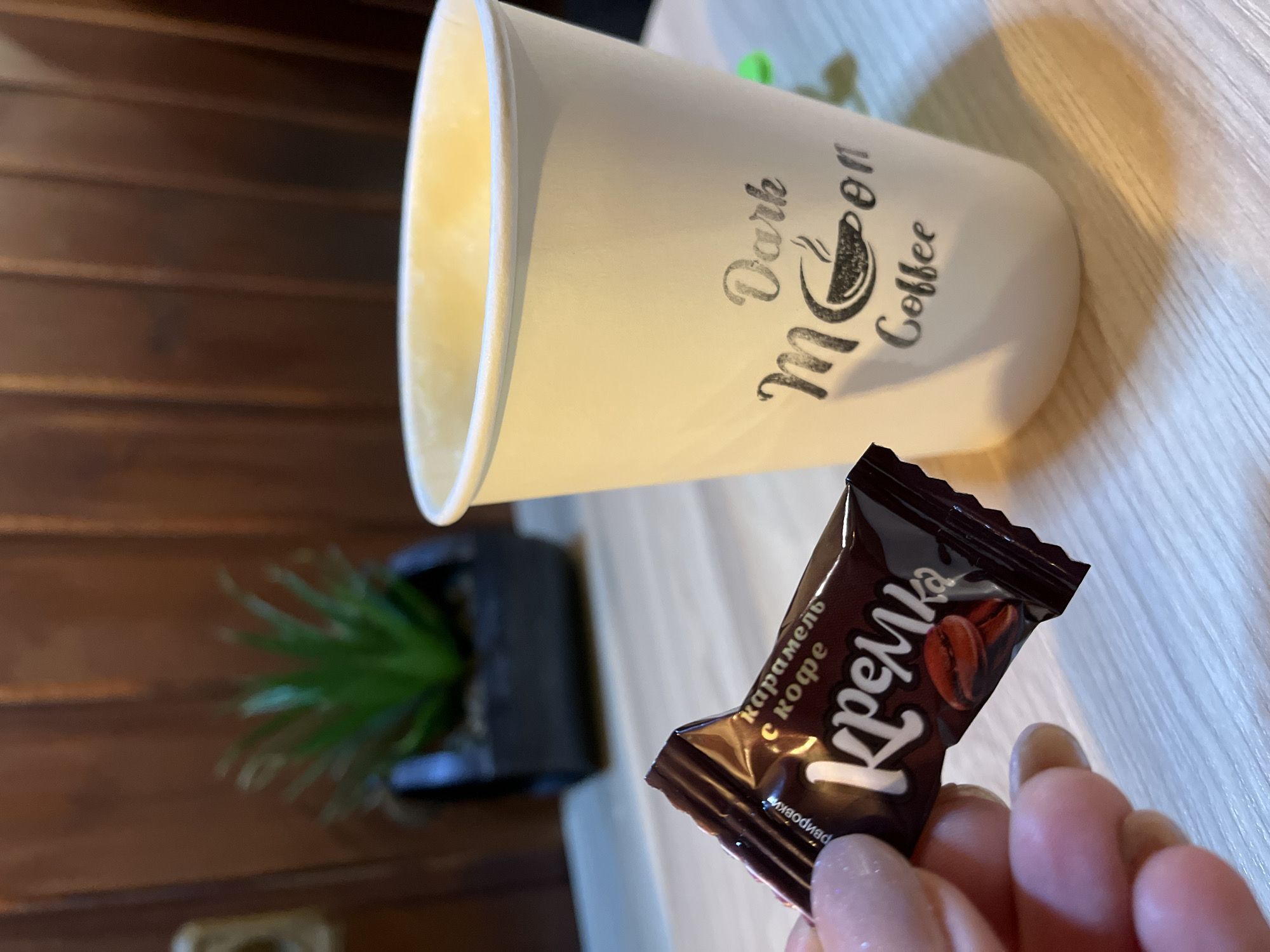 The image shows a still life composition on a wooden surface. A white paper coffee cup with "Dark Moon Coffee" printed on its side is positioned on the right. To the left of the cup is a small, dark brown wrapped candy labeled "Кремка" (Kremka) with coffee beans printed on the wrapper, held in a person's hand. In the background, a small potted plant can be seen near a dark wooden wall. The location is possibly Petropavlovsk-Kamchatsky, Russia, given the candy wrapper's language and prevalence in that region.