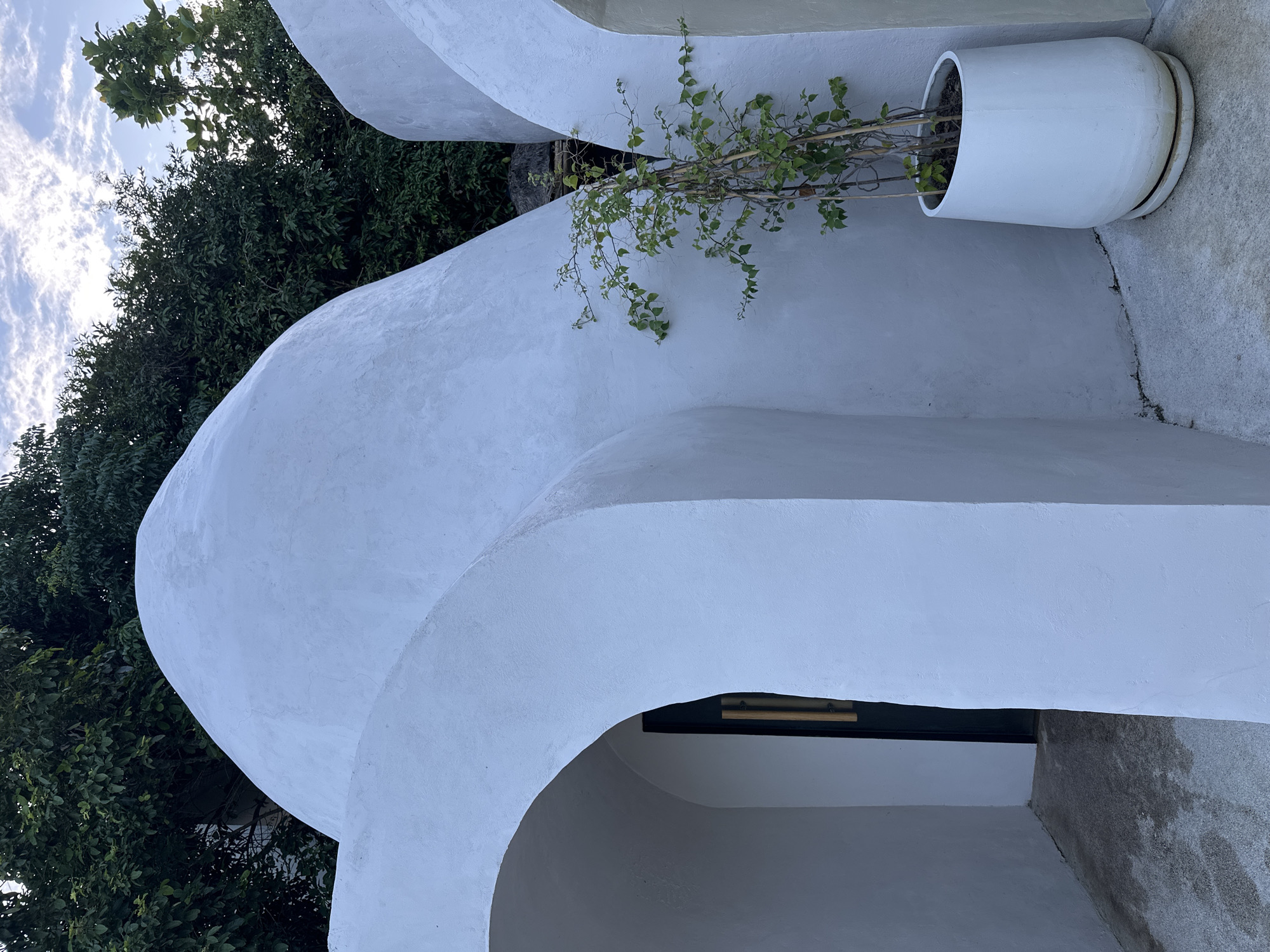 The image captures a close-up view of a white, curvilinear building structure with an arched entryway. The building has a smooth, stucco-like finish and appears to be partially submerged or built into a hillside, with lush green foliage visible behind and above it. A narrow, dark-colored door is visible through the archway. To the right of the structure, a slender green plant in a large white planter is positioned on a concrete ground surface. The overall aesthetic is modern and organic, blending with the natural surroundings. The sky above features scattered clouds. The scene is set in a tropical environment.
