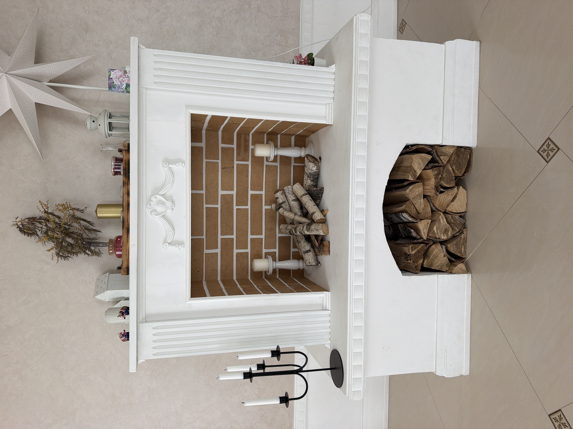 The image shows a white, decorative fireplace with a brick-lined interior. Above the fireplace opening, there is a mantelpiece adorned with various decorative items, including a branched candelabra with white candles on the left, a stack of birch logs in the fireplace, and a single white candle on a stand inside the fireplace on the right. The mantelpiece itself is decorated with carved moldings and various small objects such as figurines, a vase with dried flowers, a golden candle, a jar, a lantern, and a decorative box. A large white paper star hangs on the wall behind the mantelpiece. The floor is tiled with a beige color and a diamond pattern. The fireplace is situated against a textured, light-colored wall. The scene appears to be an indoor setting, possibly a living room, in a home. The location is specified as Дабрынёўскі сельскі Савет, Belarus.