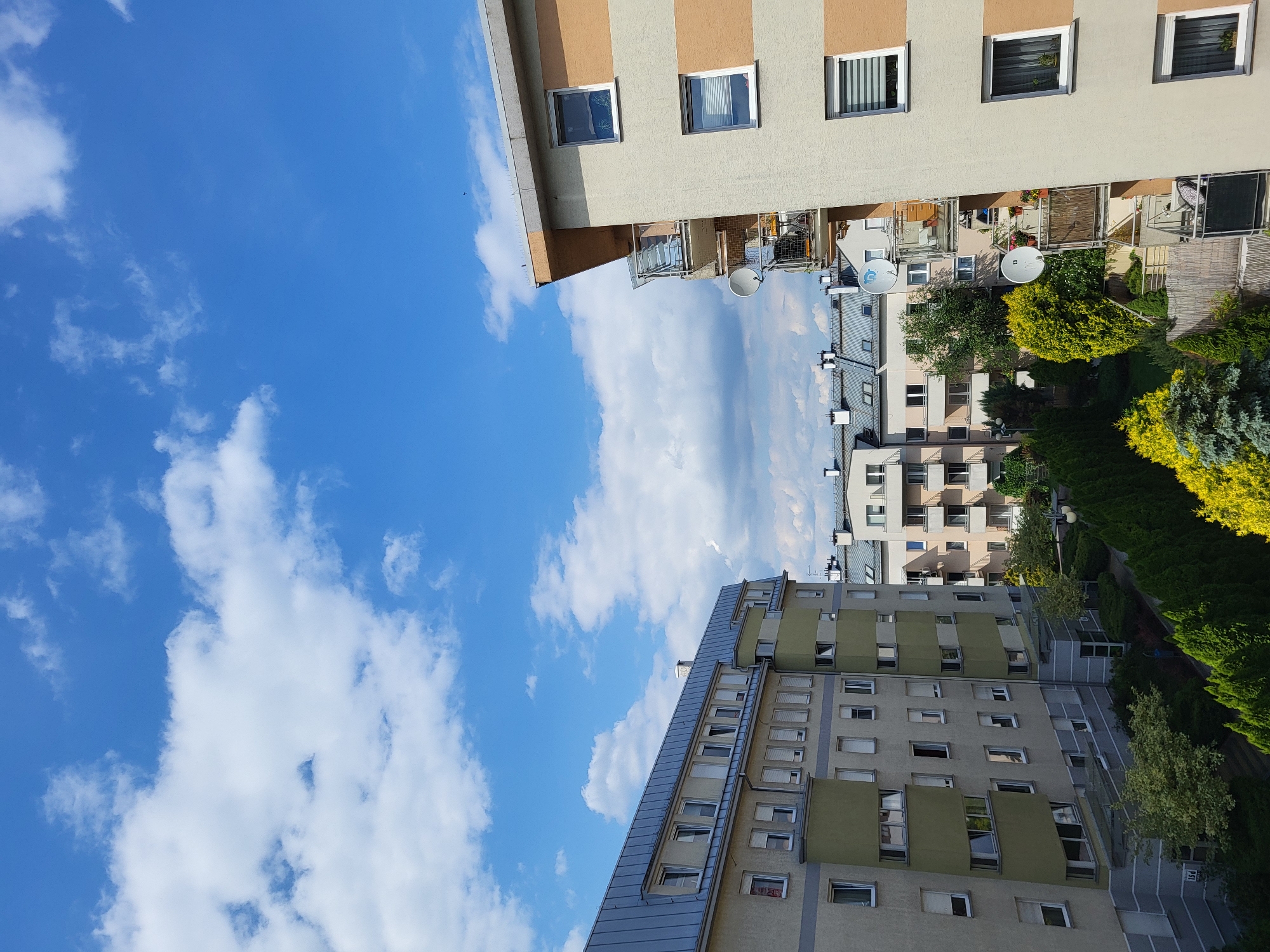 This is an outdoor, eye-level shot of apartment buildings under a bright, partly cloudy sky. The sky is a vibrant blue with scattered white cumulus clouds. On the left, a multi-story apartment building with a gray, sloped metal roof is visible. Its facade is a pale yellow with green sections and numerous windows. To the right, another apartment building with a beige and orange striped facade features balconies with satellite dishes and railings. In the background, more apartment buildings of varying heights and architectural styles are visible. A line of evergreen trees and some deciduous trees with yellow foliage are situated in the foreground. The scene appears to be a residential area within a city.