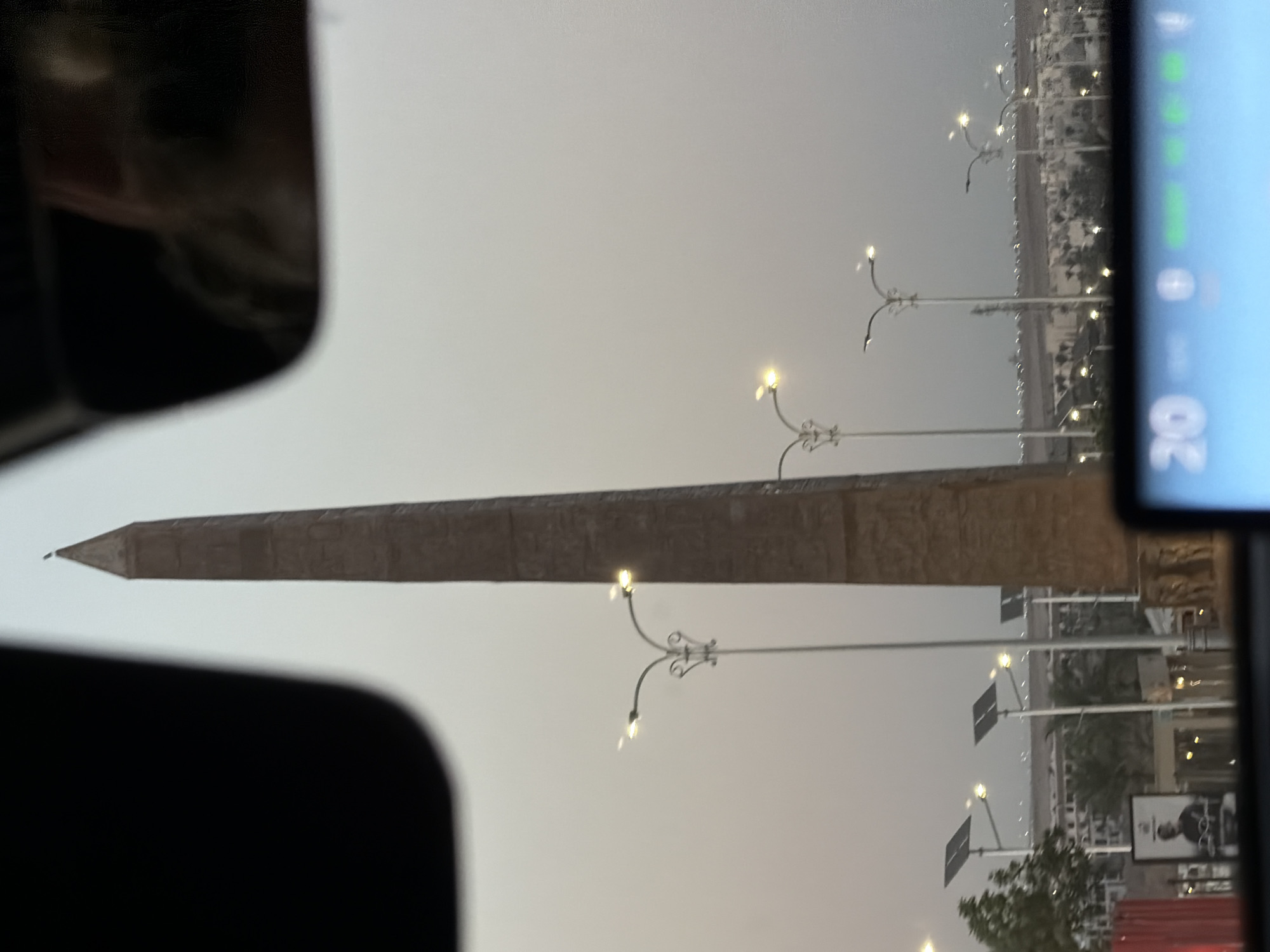 A tall, ancient Egyptian obelisk stands prominently in the center of the frame, its weathered stone surface etched with hieroglyphics. The sky behind it is a hazy, pale gray, suggesting either dawn or dusk. Several ornate streetlights with multiple bulbs dot the landscape, some illuminated, casting a warm glow. In the background, a row of modern buildings and palm trees are visible, hinting at an urban environment. Solar panels are mounted on poles, further indicating a developed area. The foreground is partially obscured by the dark interior of a vehicle, with a digital display showing "20" and other indicators. The reflection of a person's face is visible in the upper right corner, cast on a dark surface, likely a car's rearview mirror. The overall scene appears to be a street in Sharm El Sheikh, Egypt, capturing a blend of ancient history and modern life.